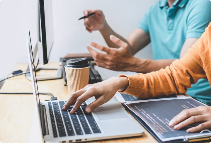 Two people working at a desk with laptops and a desktop monitor discuss sourcing spare parts for a gas turbine. One types while the other gestures with a pen. A coffee cup and open notebook complete the busy workspace.