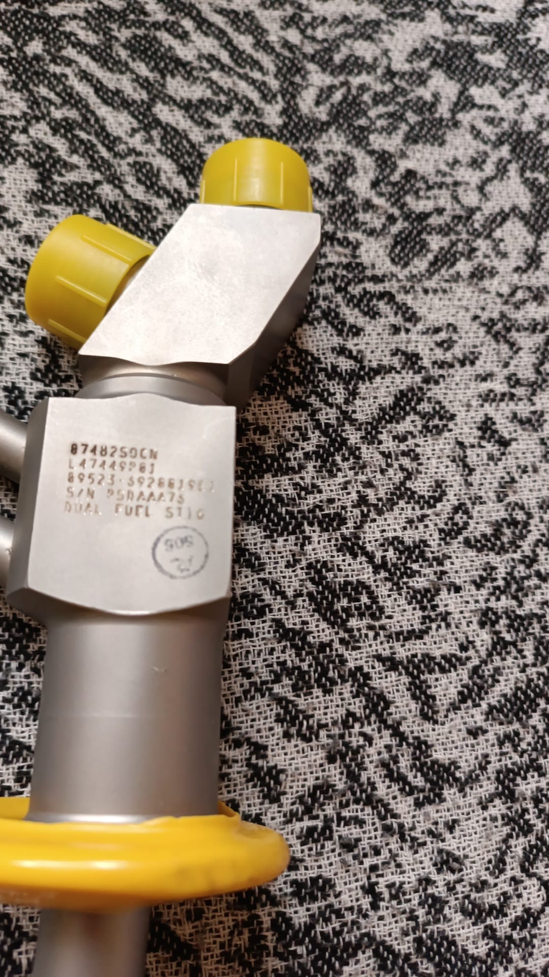 A close-up of a metal mechanical part with engraved text and yellow plastic caps, placed on a black and white patterned fabric background, showcases one of the 4 Overhauled OH Dual Fuel DF Nozzles L47449P01.