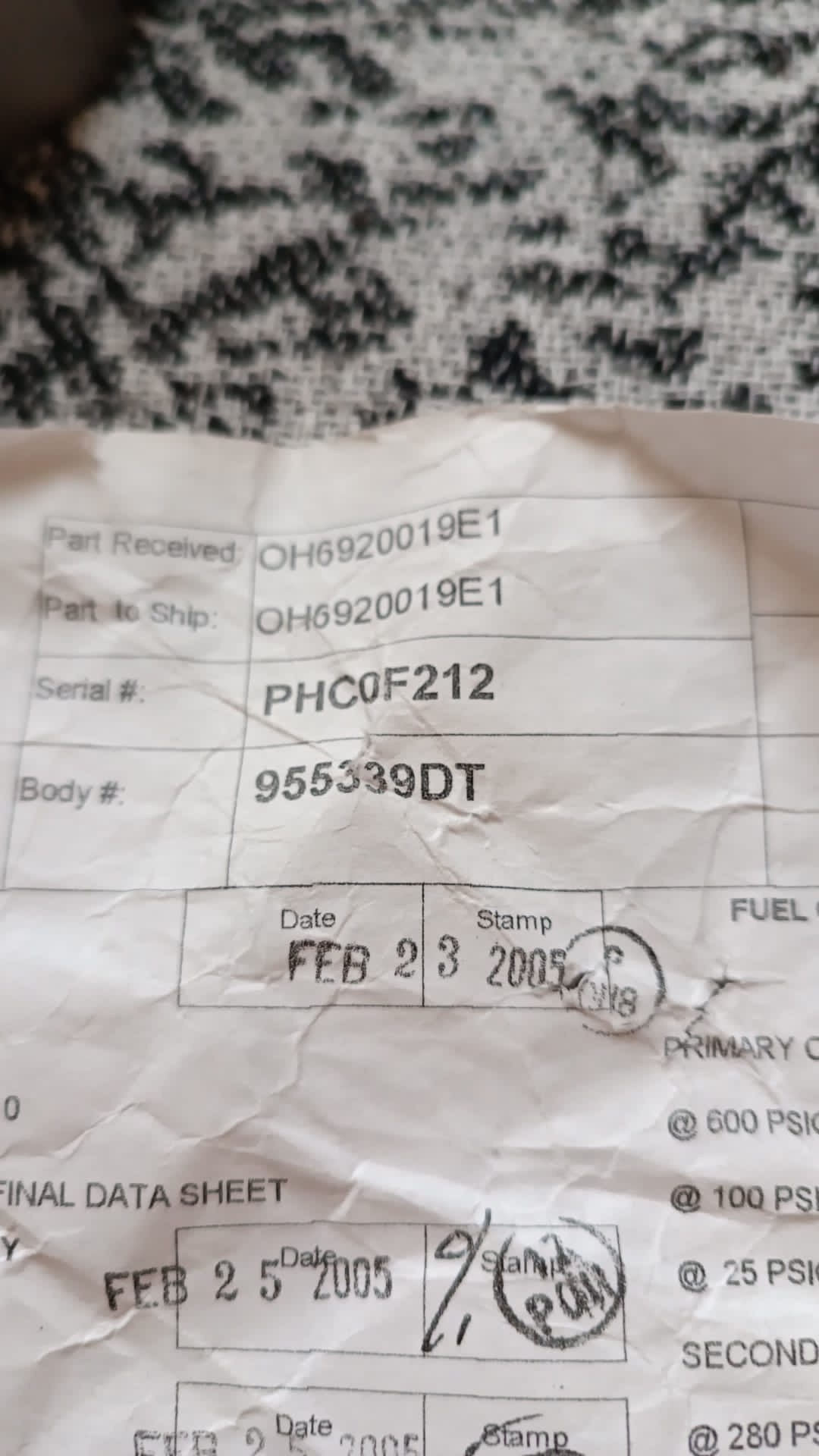 A close-up of a wrinkled paper form with printed part and serial numbers for 4 Overhauled OH Dual Fuel DF Nozzles L47449P01, plus two visible date stamps—FEB 23 2005 and FEB 25 2005. The form rests on a textured surface.