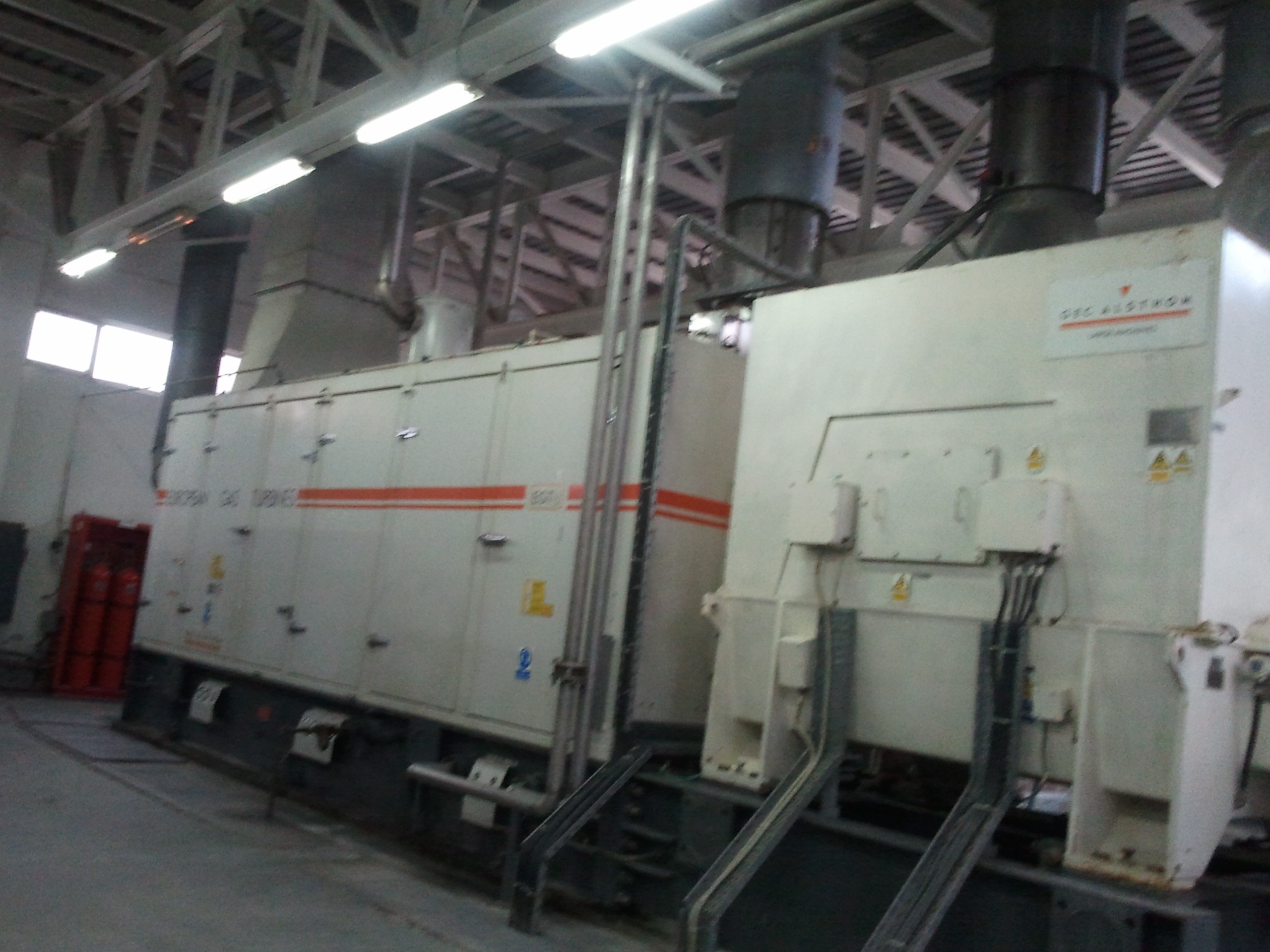 Large industrial machines such as the Siemens SGT 200 Gas Turbine Packages, equipped with pipes and control panels, stand inside a factory under fluorescent lighting. Warning signs and labels are visible on the equipment, alongside shelves stocked with essential spare part supplies nearby.