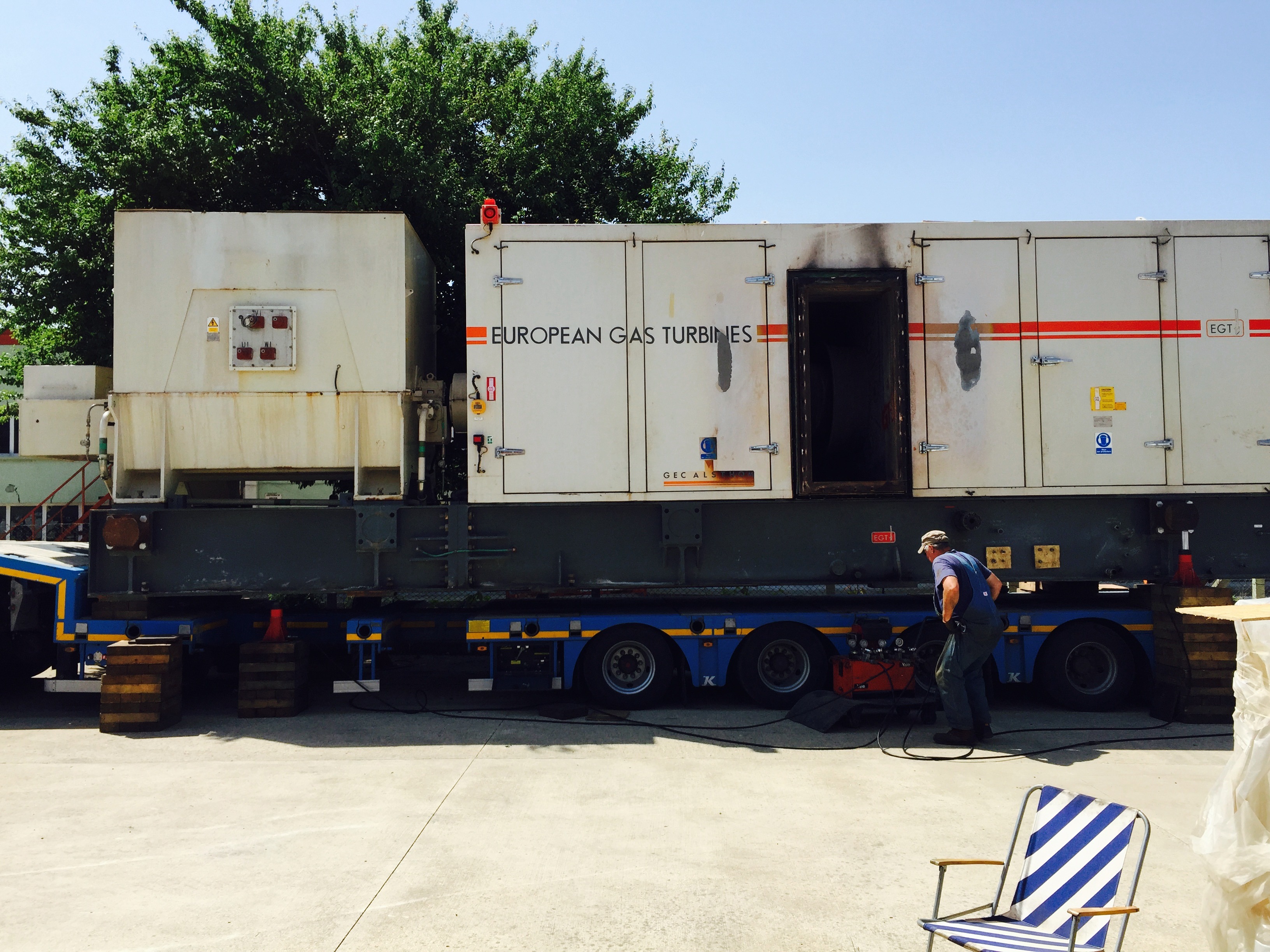 A worker stands near a large Siemens SGT 200 Gas Turbine Package on a trailer, highlighting the scale of heavy industry at work. The equipment features open panels and cables, with wooden blocks securing the trailer wheels. Nearby, a striped chair sits on the pavement.