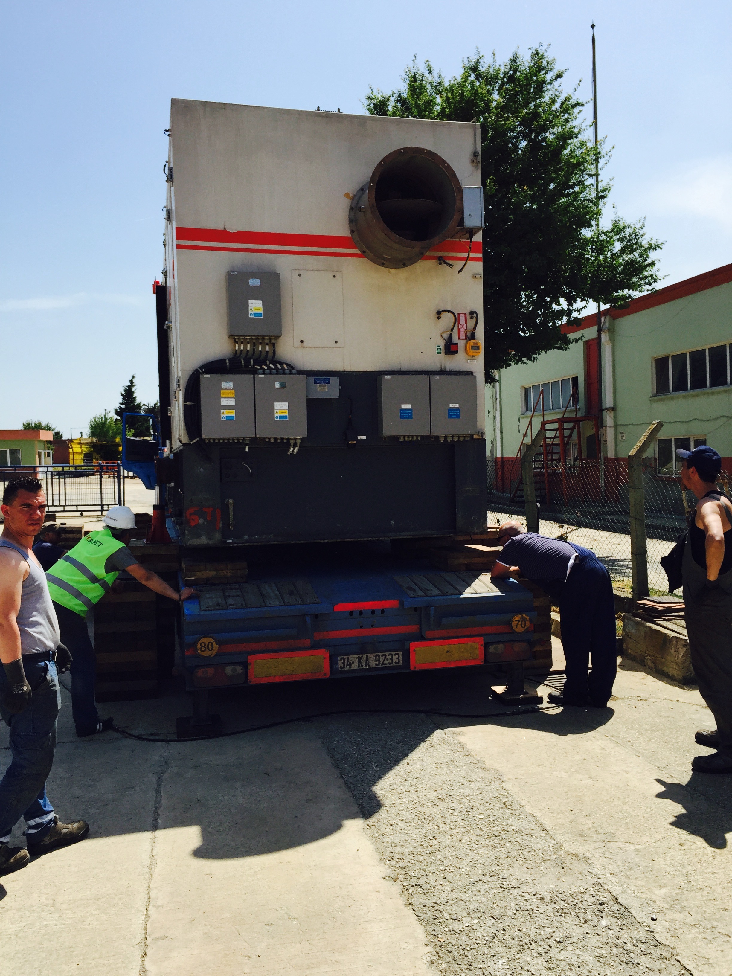 Workers stand around a large Siemens SGT 200 Gas Turbine Package being loaded onto a flatbed truck outside, near industrial buildings, on a sunny day. This impressive display of heavy industry highlights the coordination and expertise required to transport such advanced Siemens equipment.