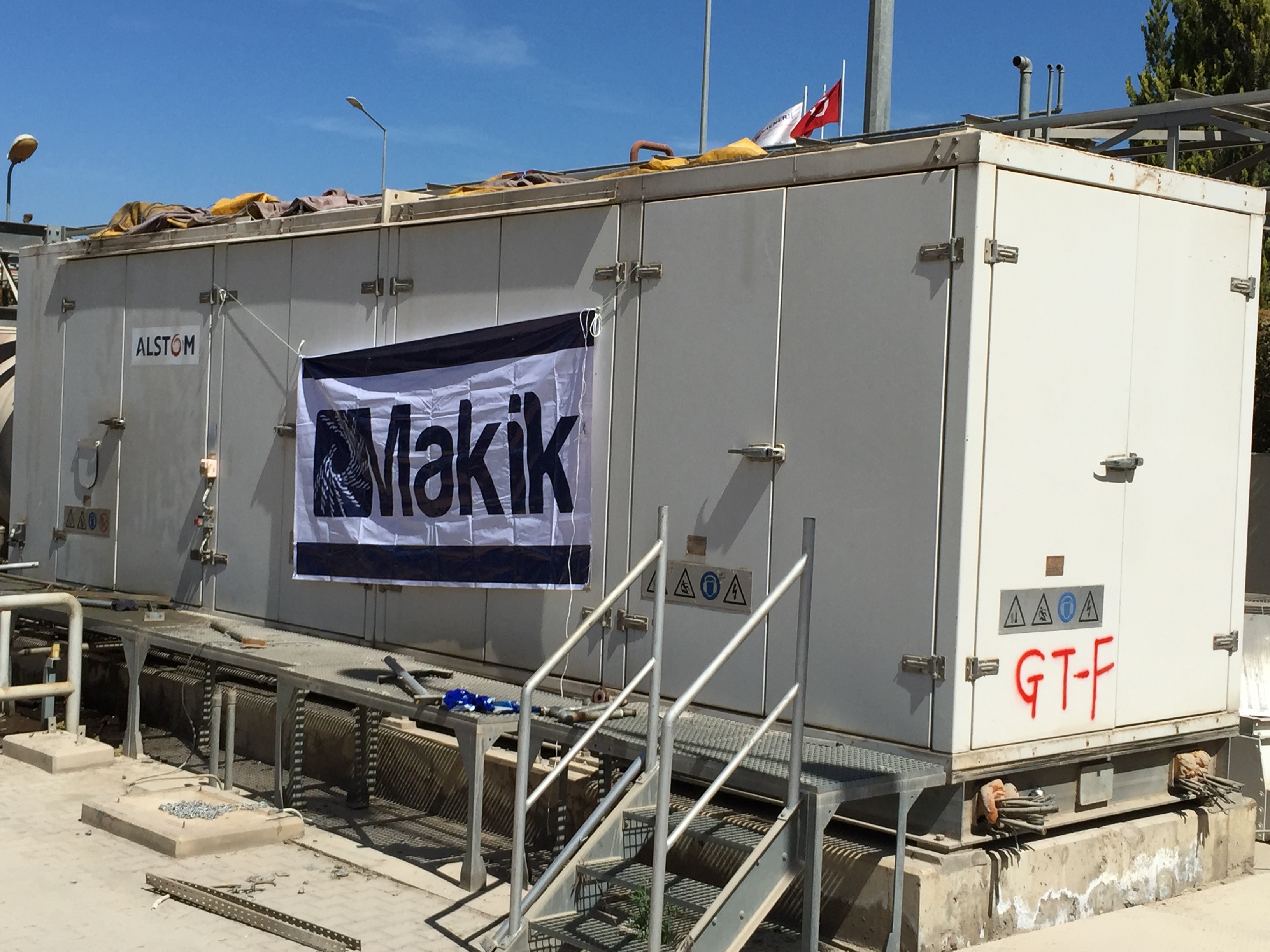 Six large white industrial Siemens SGT 100 gas turbine packages, designed for heavy industry applications, are showcased with a Makik banner and ALSTOM logo. Each container features stairs, warning signs, and is elevated on a platform with railings. Visible in the background are pipes and a flag beneath a blue sky—these units are ideal for housing spare part components such as the LM2500.