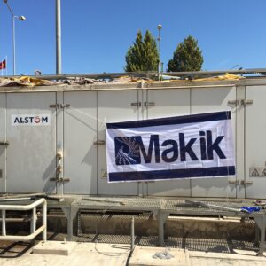 A large white refurbished industrial unit with ALSTOM and Makik logos, pipes, and safety signs under a clear blue sky, possibly housing sold 6 sets of Siemens SGT 100 gas turbine packages.