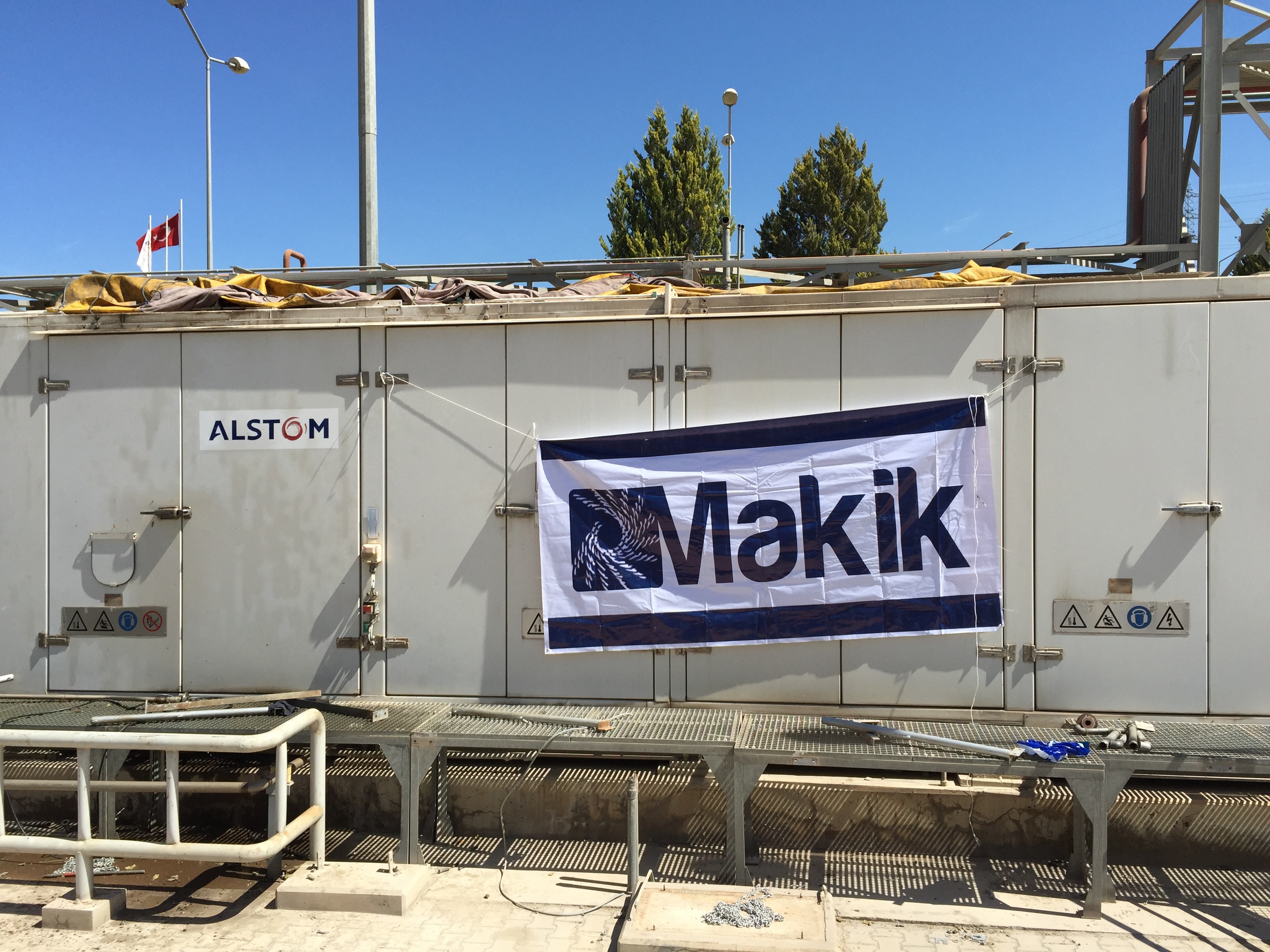 A large white refurbished industrial unit with ALSTOM and Makik logos, pipes, and safety signs under a clear blue sky, possibly housing sold 6 sets of Siemens SGT 100 gas turbine packages.