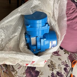 A used blue SOLD noah proportional electrical actuator valve delivery, partially bubble-wrapped, rests on a floral-patterned sofa with a purple pillow nearby. An orange-handled tool lies on the right, suggesting turbine system components.