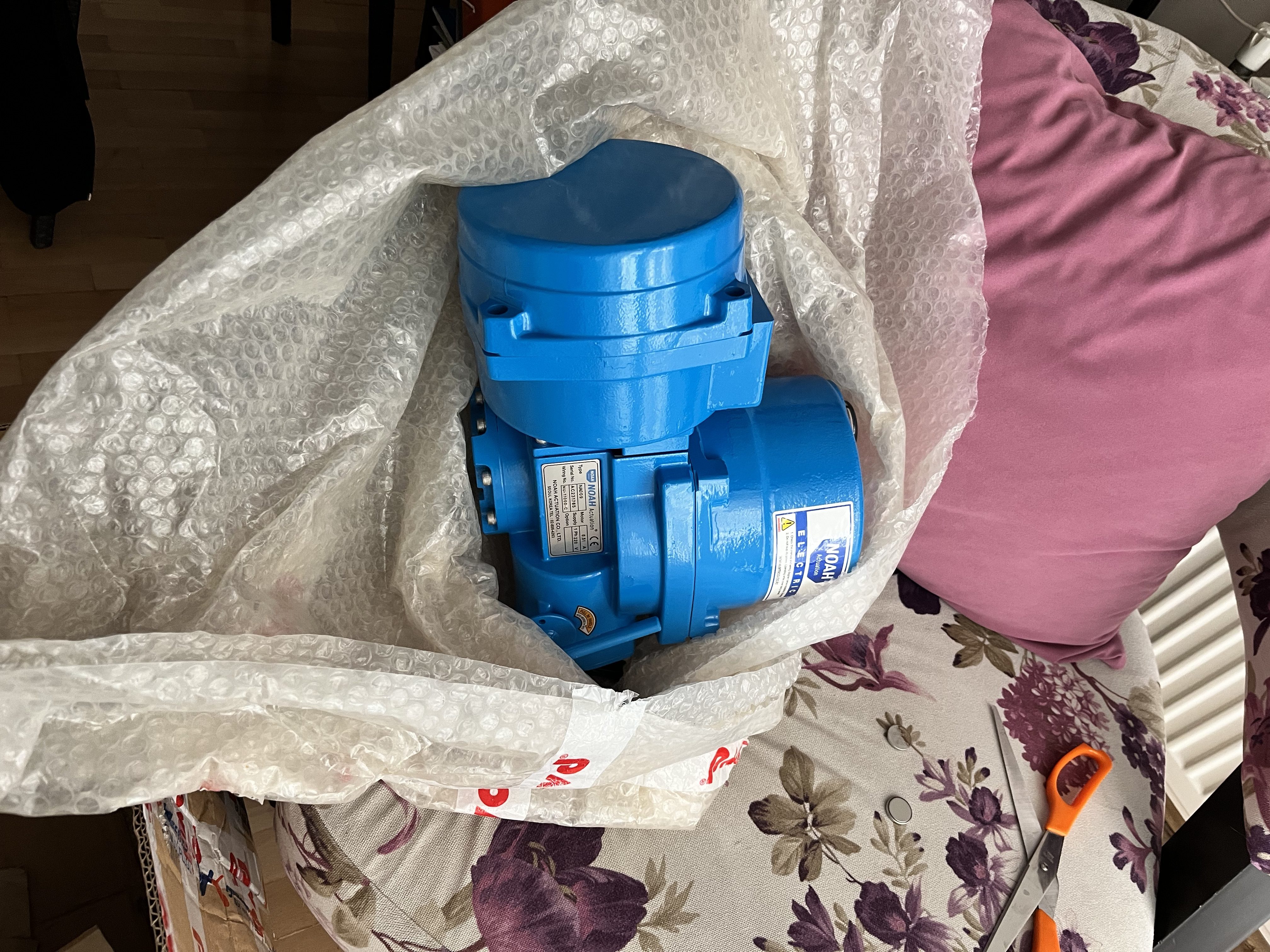 A used blue SOLD noah proportional electrical actuator valve delivery, partially bubble-wrapped, rests on a floral-patterned sofa with a purple pillow nearby. An orange-handled tool lies on the right, suggesting turbine system components.