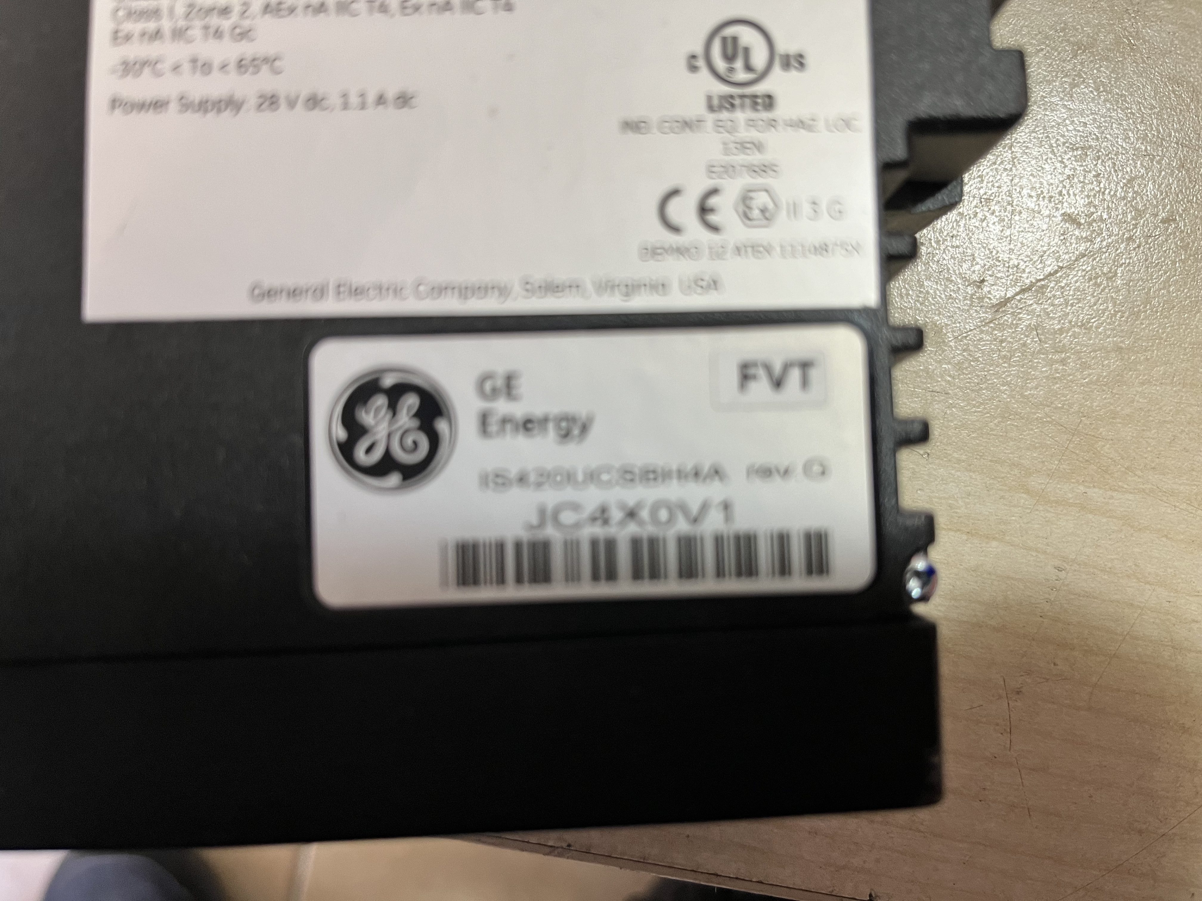 Close-up photo of a Used GE Energy IS420UCSBH4A UCSB CONTROLLER MODULE label, revision C, serial JC4X0V1 with certification marks, attached to a black electronic unit on a light surface.