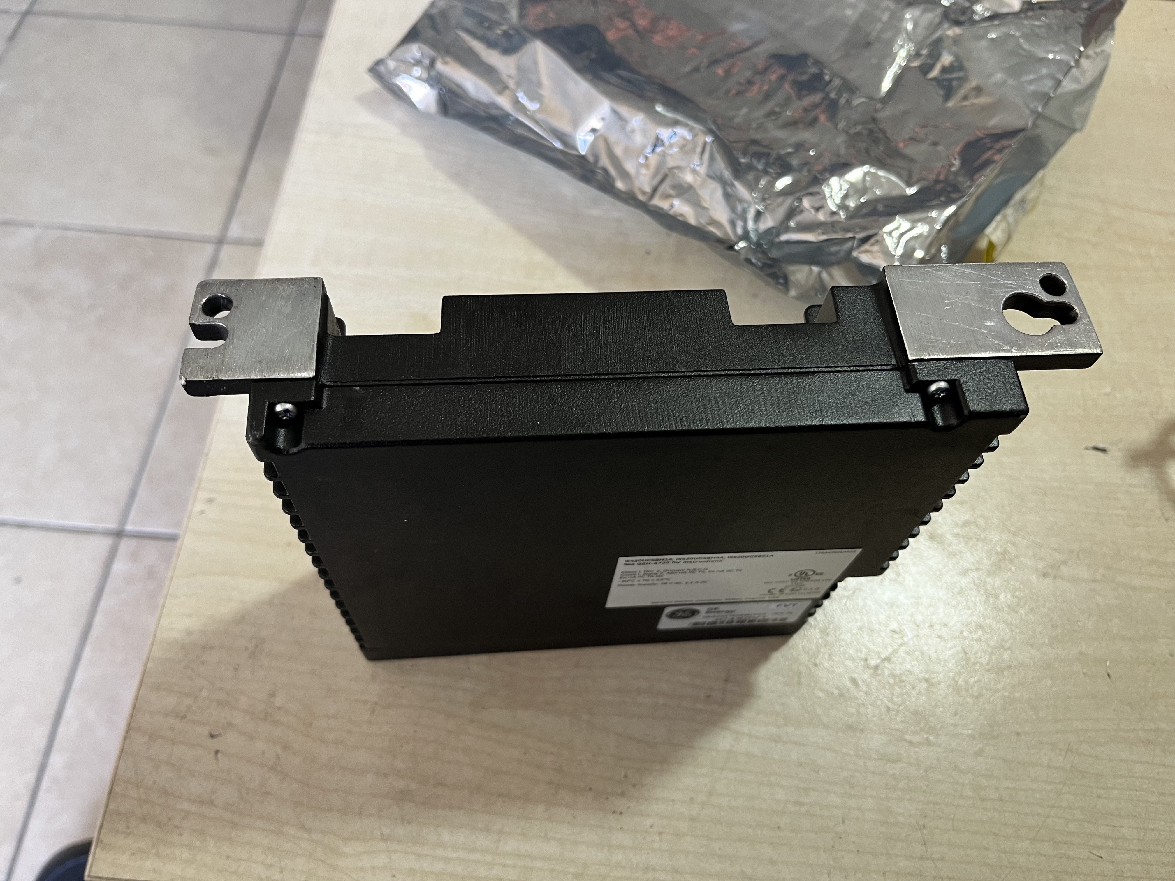 A Used GE Energy IS420UCSBH4A UCSB CONTROLLER MODULE with metal mounting brackets on both ends rests on a light desk beside a crumpled silver anti-static bag, ready for use as a spare part.
