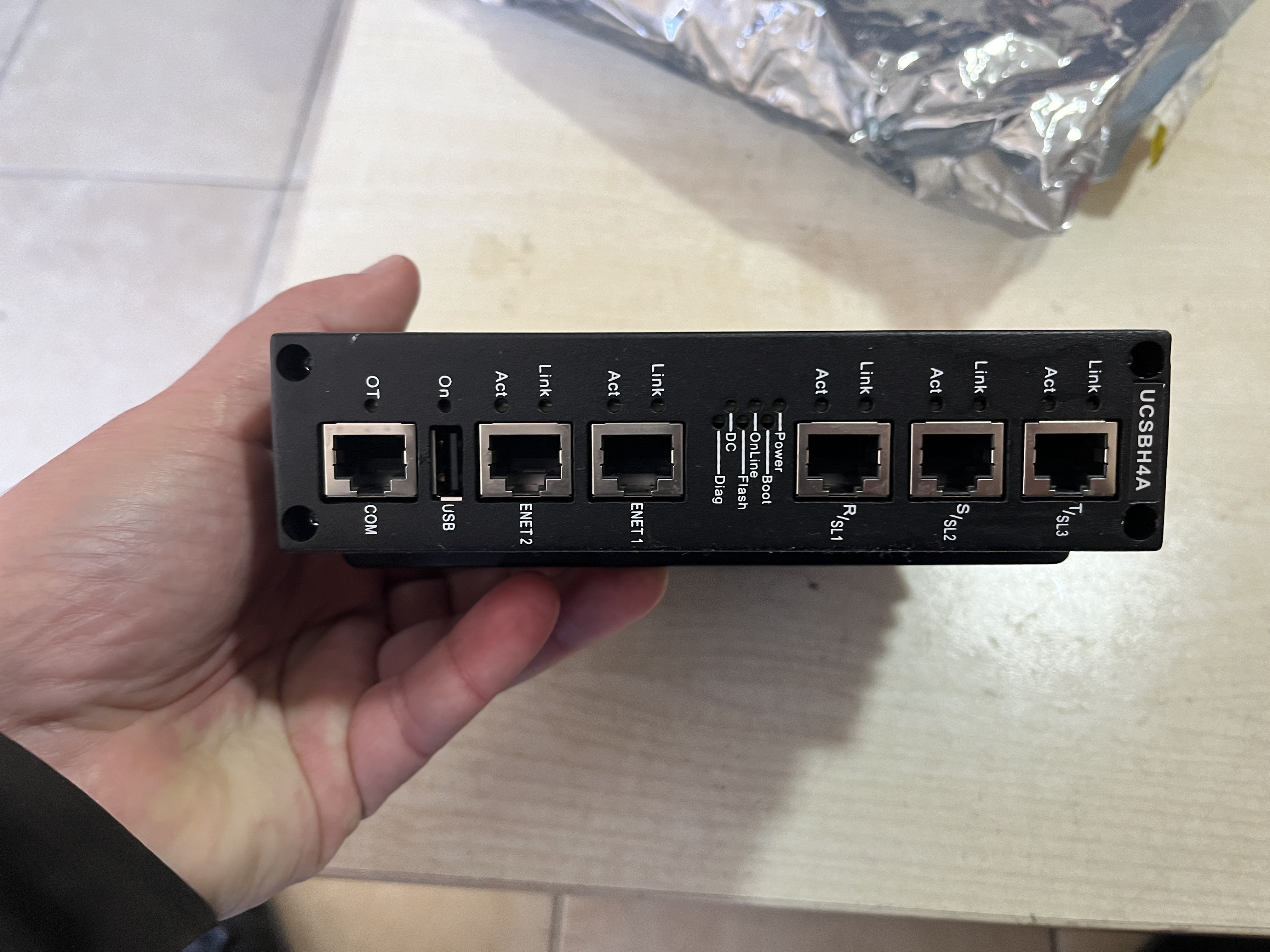 A hand holds a used GE Energy IS420UCSBH4A UCSB Controller Module with labeled Ethernet ports, resting on a light-colored surface.
