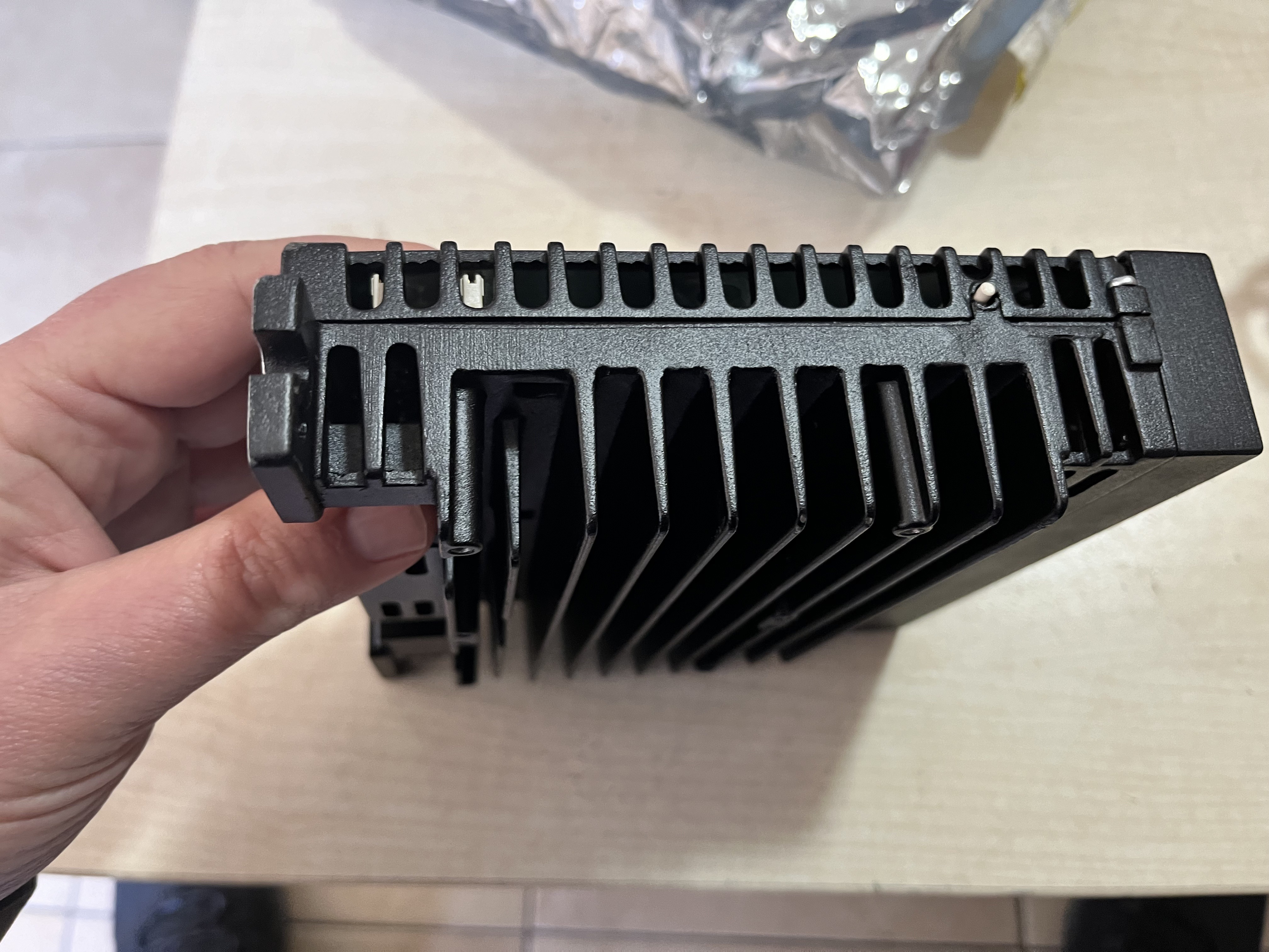 A hand holds a used GE Energy IS420UCSBH4A UCSB Controller Module, black and rectangular with large cooling fins and vents, viewed from the top over a light-colored surface.