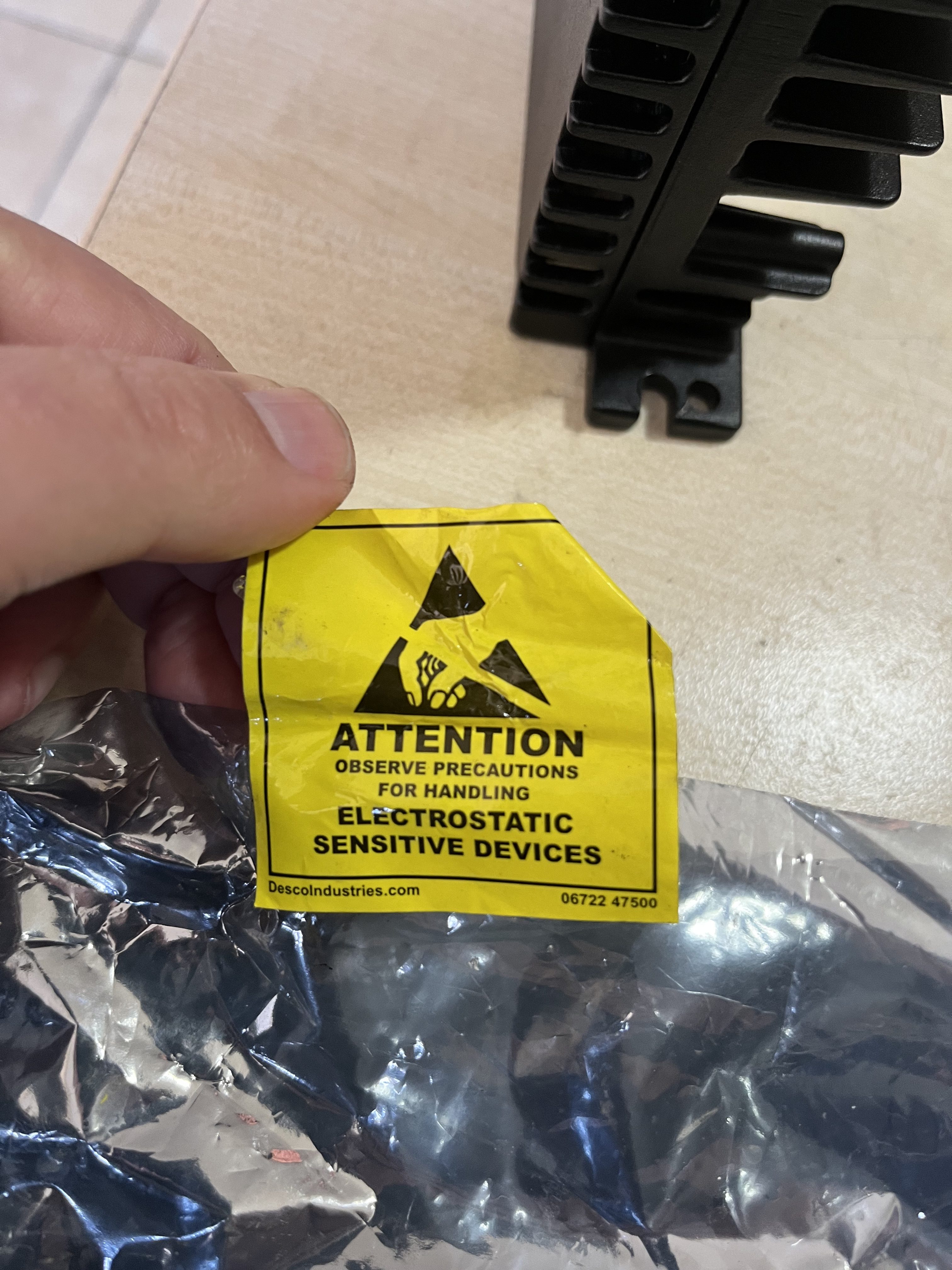 A hand holds a crumpled yellow caution label reading “Attention: Observe precautions for handling electrostatic sensitive devices” above a metallic anti-static bag containing a black Used GE Energy IS420UCSBH4A UCSB CONTROLLER MODULE.