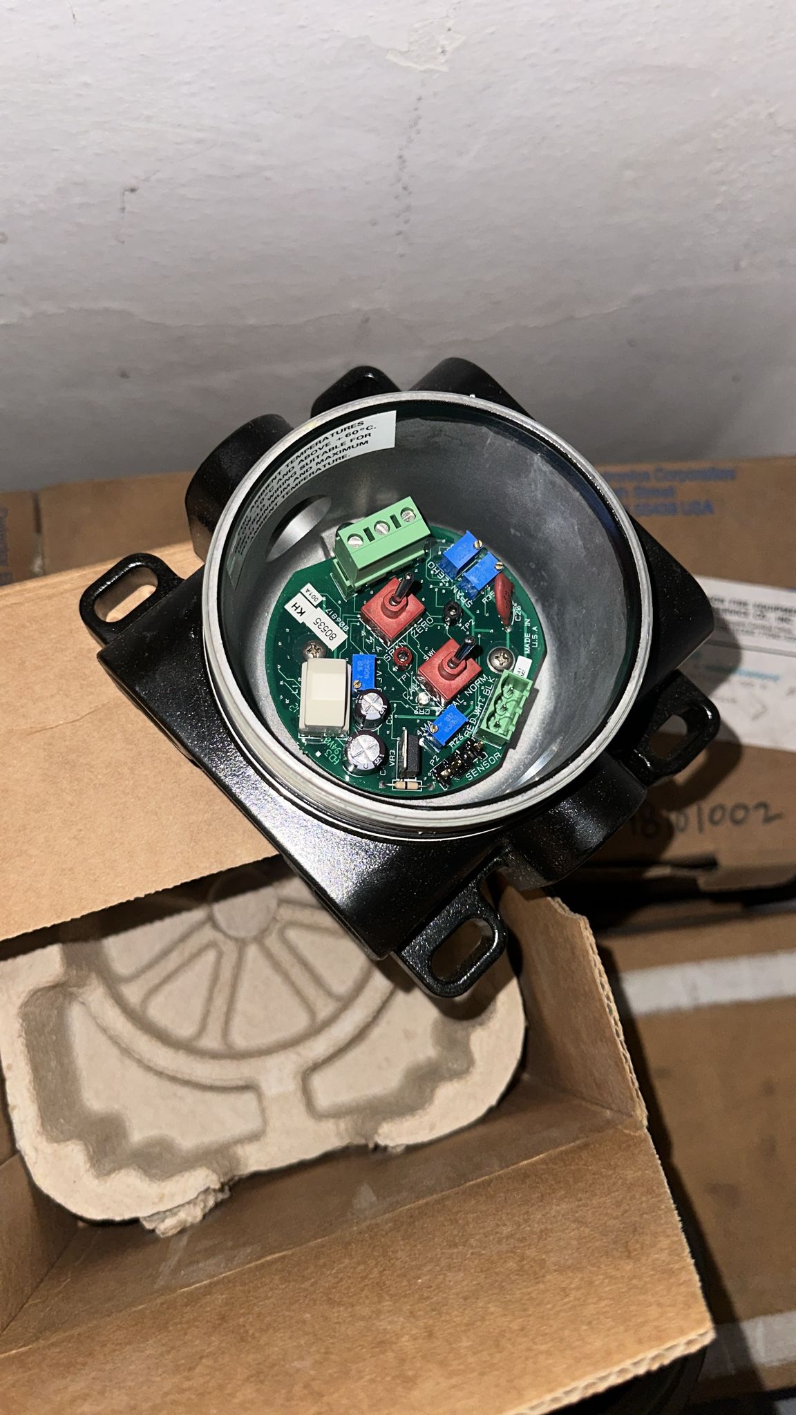 A close-up of an opened Det-tronics STB1A2MW Model STB Sensor Termination Boxes Separation Kit, revealing the internal circuit board with various components, resembling a spare part commonly used in heavy industry applications, placed inside a cardboard box with protective packaging material underneath.