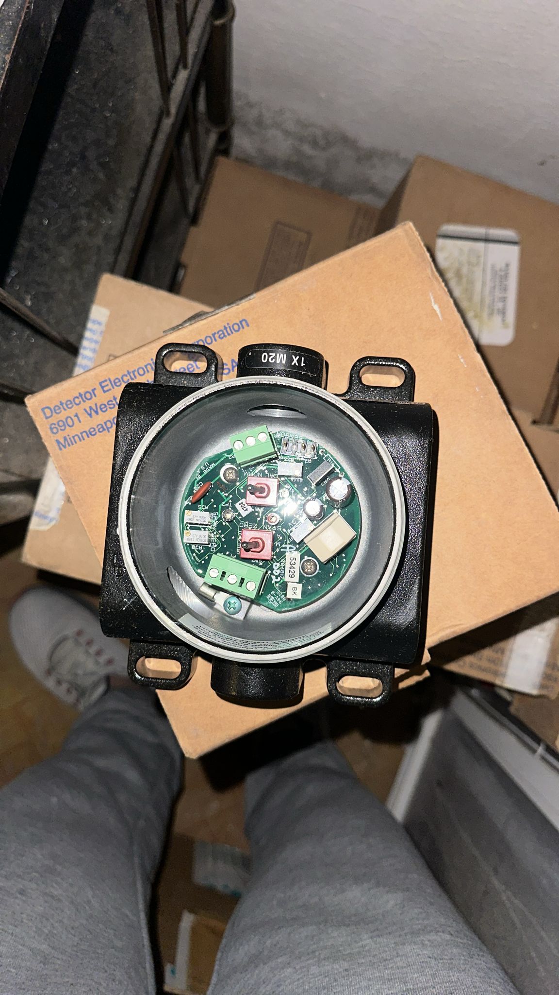 A close-up of an opened Det-tronics STB1A2MW Model STB Sensor Termination Boxes Separation Kit, showing its internal circuit board, held above cardboard boxes and a person’s legs in gray pants, viewed from above.