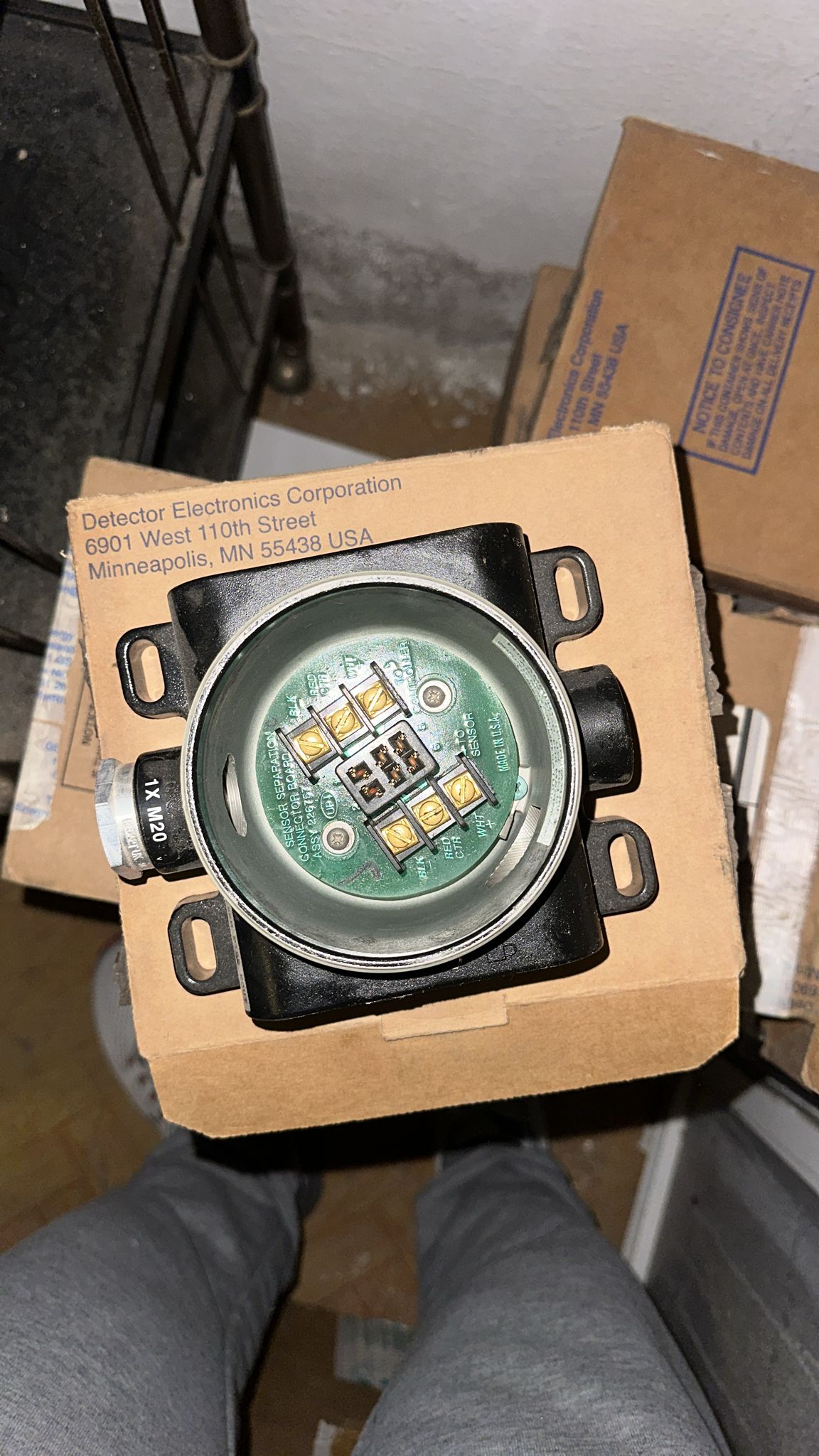 A close-up of a Det-tronics STB1A2MW Model STB Sensor Termination Boxes Separation Kit with its electronic sensor components and exposed circuit board, placed on a cardboard box labeled Detector Electronics Corporation, Minneapolis, MN. Designed for heavy industry applications such as LM6000 and LM2500 gas turbines, the kit is surrounded by other cardboard boxes and part of a metal shelf appears in the background.
