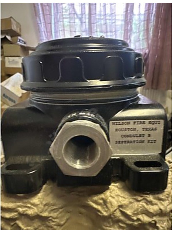 A close-up of a black mechanical device featuring a large bolt and a metal label, identified as the Det-tronics STB1A2MW Model STB Sensor Termination Boxes Separation Kit, typically found in heavy industry settings. Boxes and a window can be seen in the background.