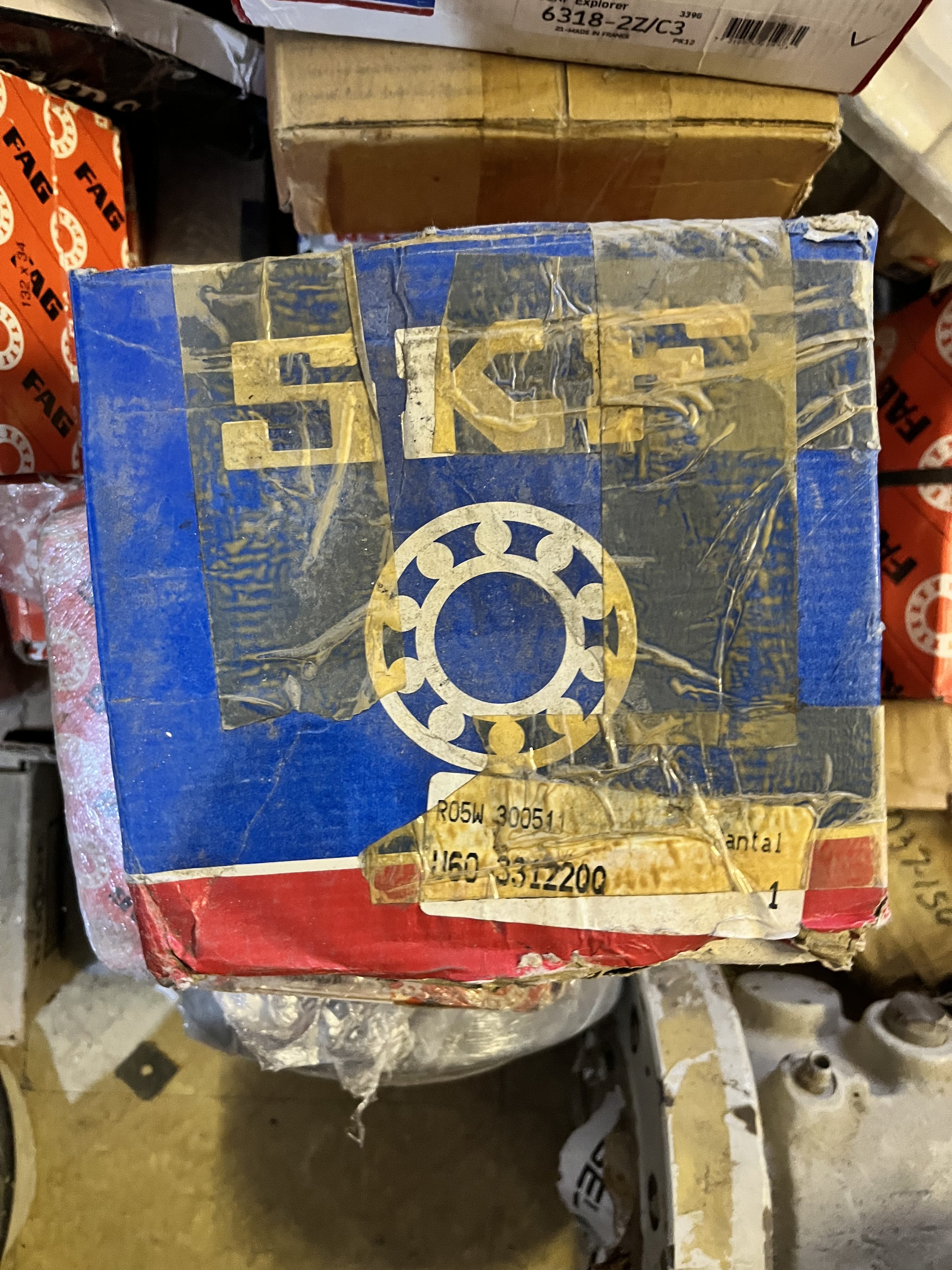 A close-up of a worn, taped SKF bearing box, mainly blue and red with a large yellow SKF logo and bearing graphic. The used box sits among other spare parts and packages in a cluttered workshop setting.