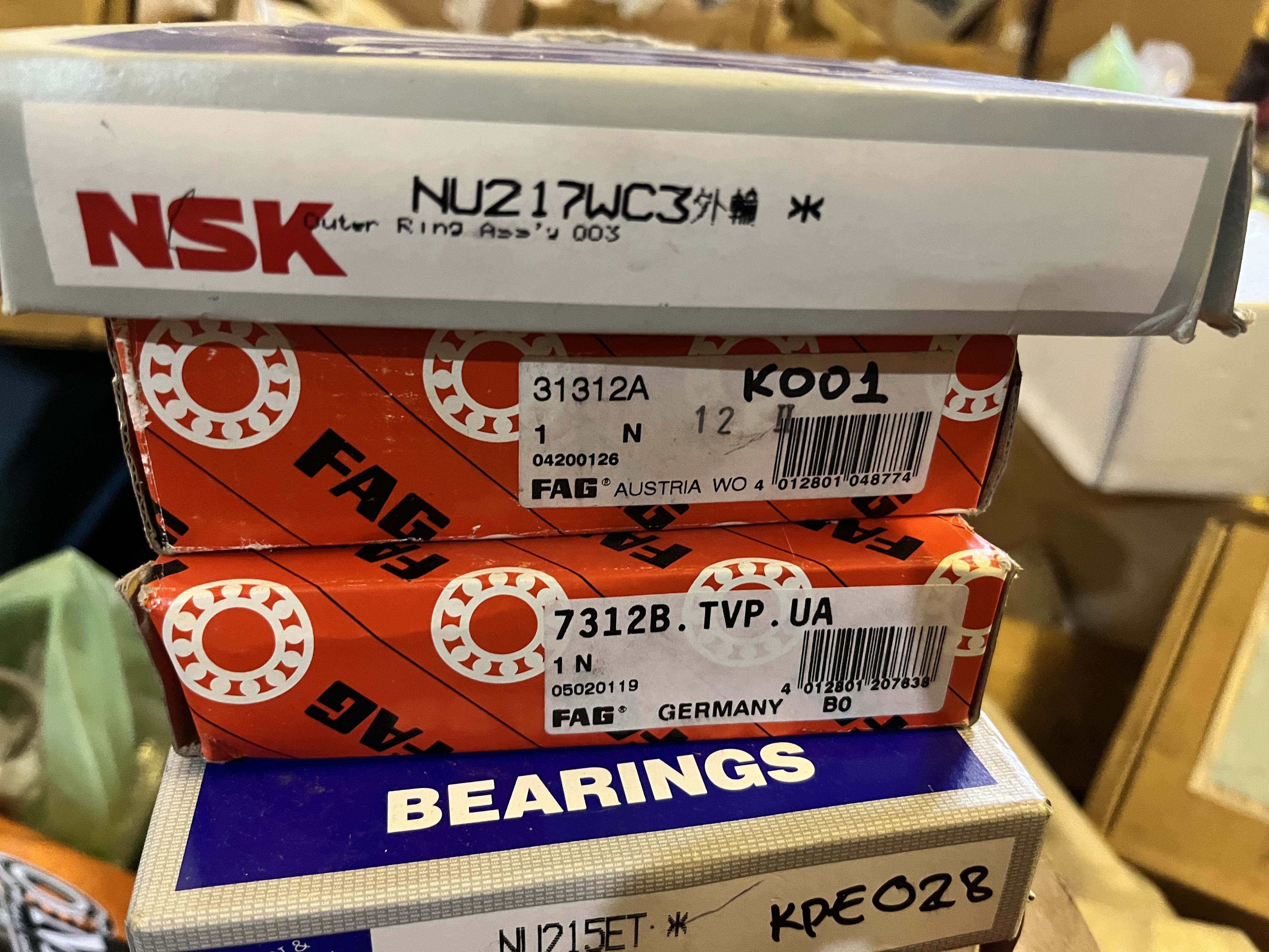A close-up of stacked bearing boxes, including obsolete and refurbished parts from brands like NSK and FAG, labeled with product numbers. The boxes sit in a workshop or storage area, with tools and other spare parts blurred in the background.