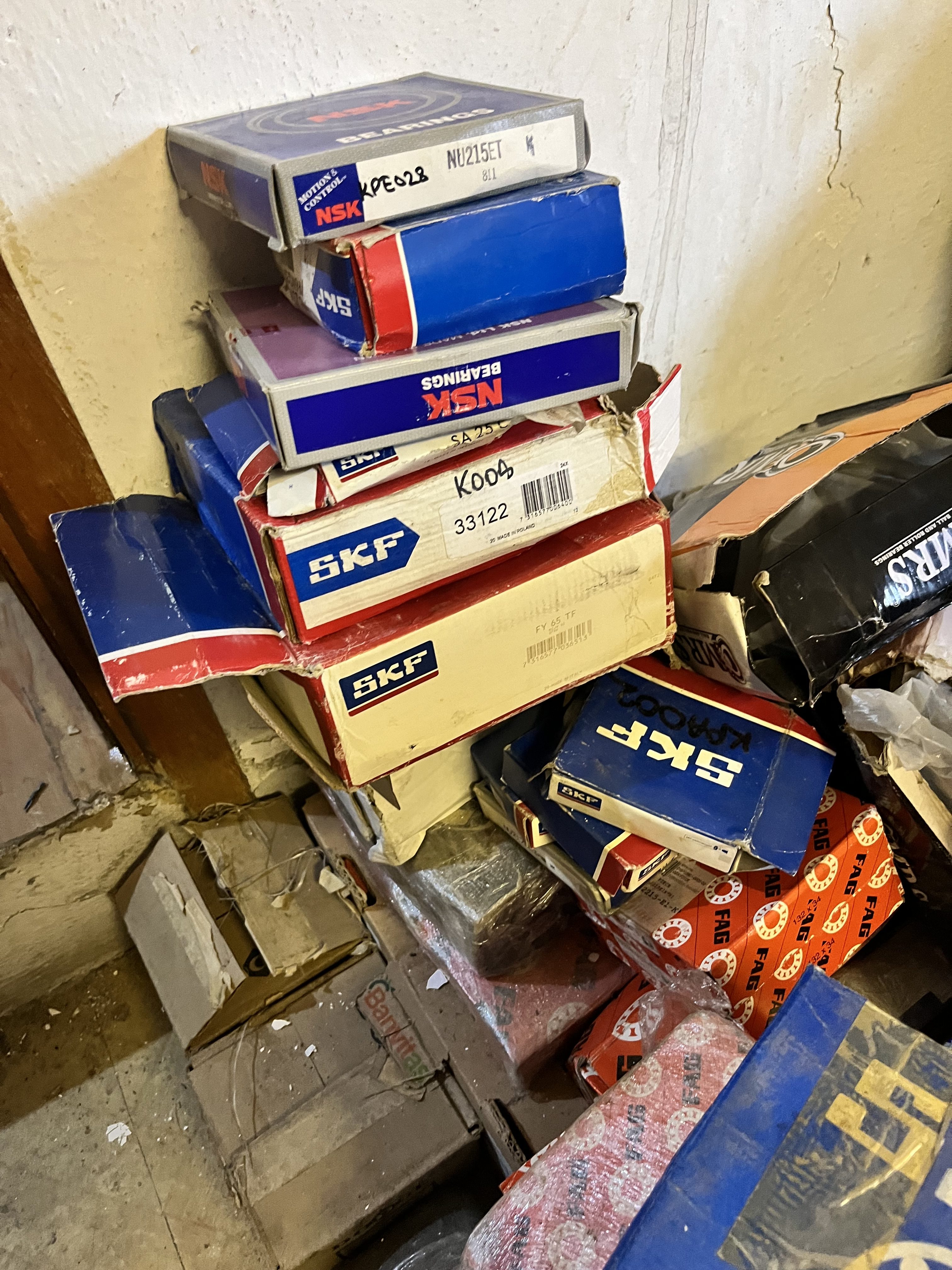 A stack of assorted boxed bearings, including some obsolete NSK and SKF models, is piled against a wall in a cluttered storage space—possibly spare parts for equipment like the LM6000 gas turbine—amid various other boxes and items.