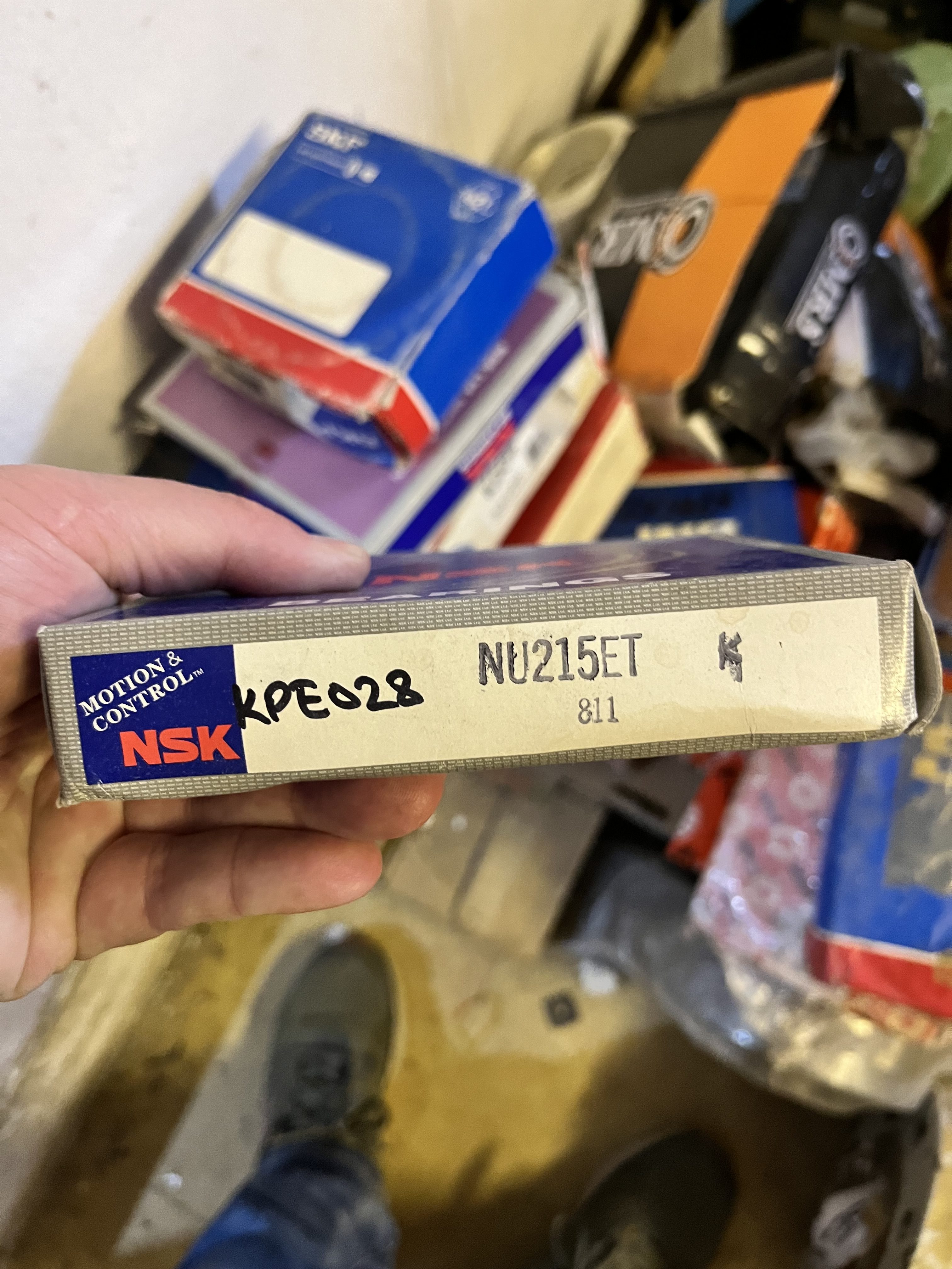 A hand holds a box labeled NSK NU215ET marked KPE028, an obsolete spare part. Several other bearing boxes are stacked in the background on the floor, with a persons shoes partially visible.
