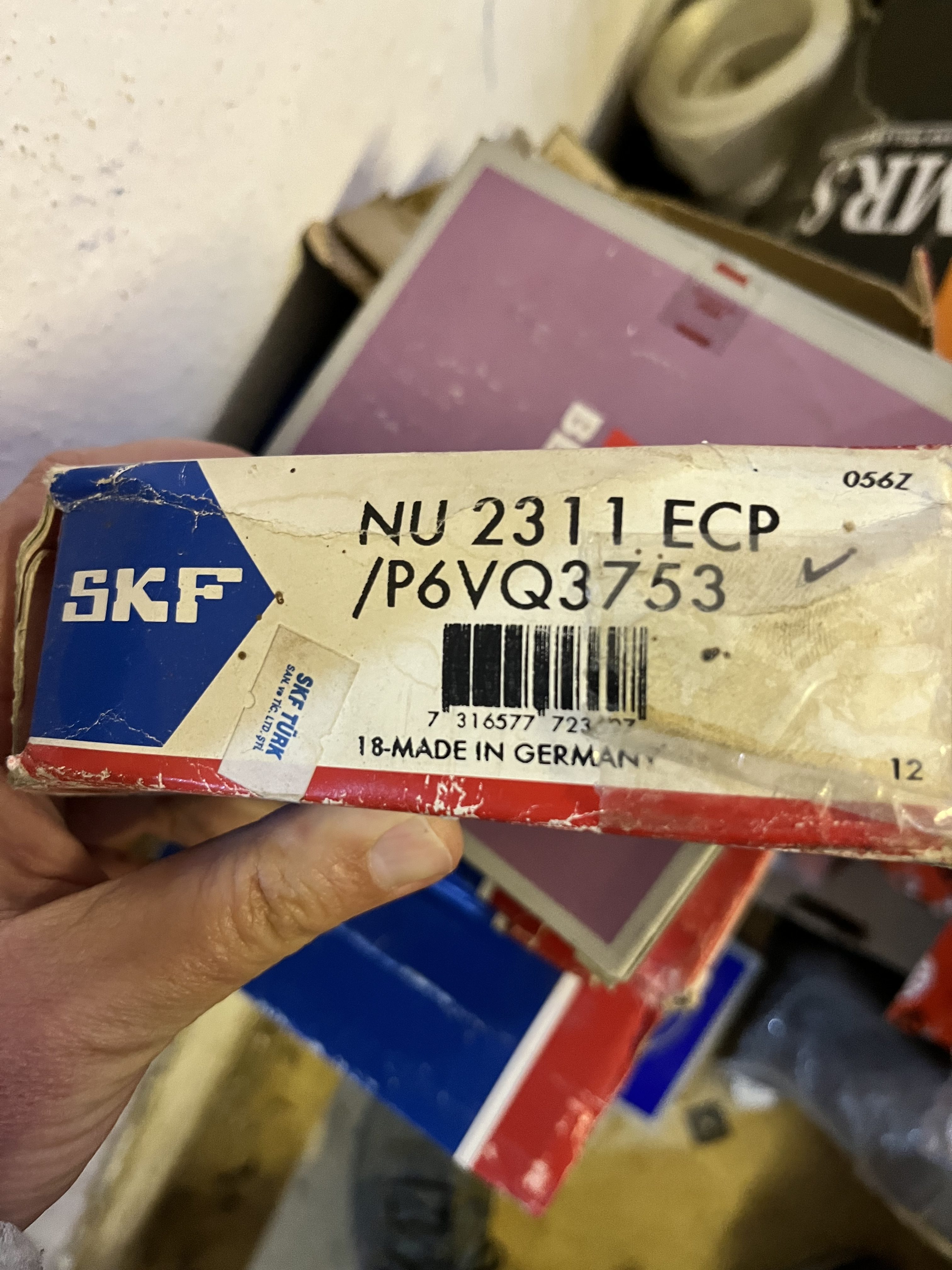 A hand holds an SKF branded box labeled NU 2311 ECP /P6VQ3753, marked MADE IN GERMANY. The worn box, possibly containing obsolete spare parts, sits among other packages with a visible barcode.