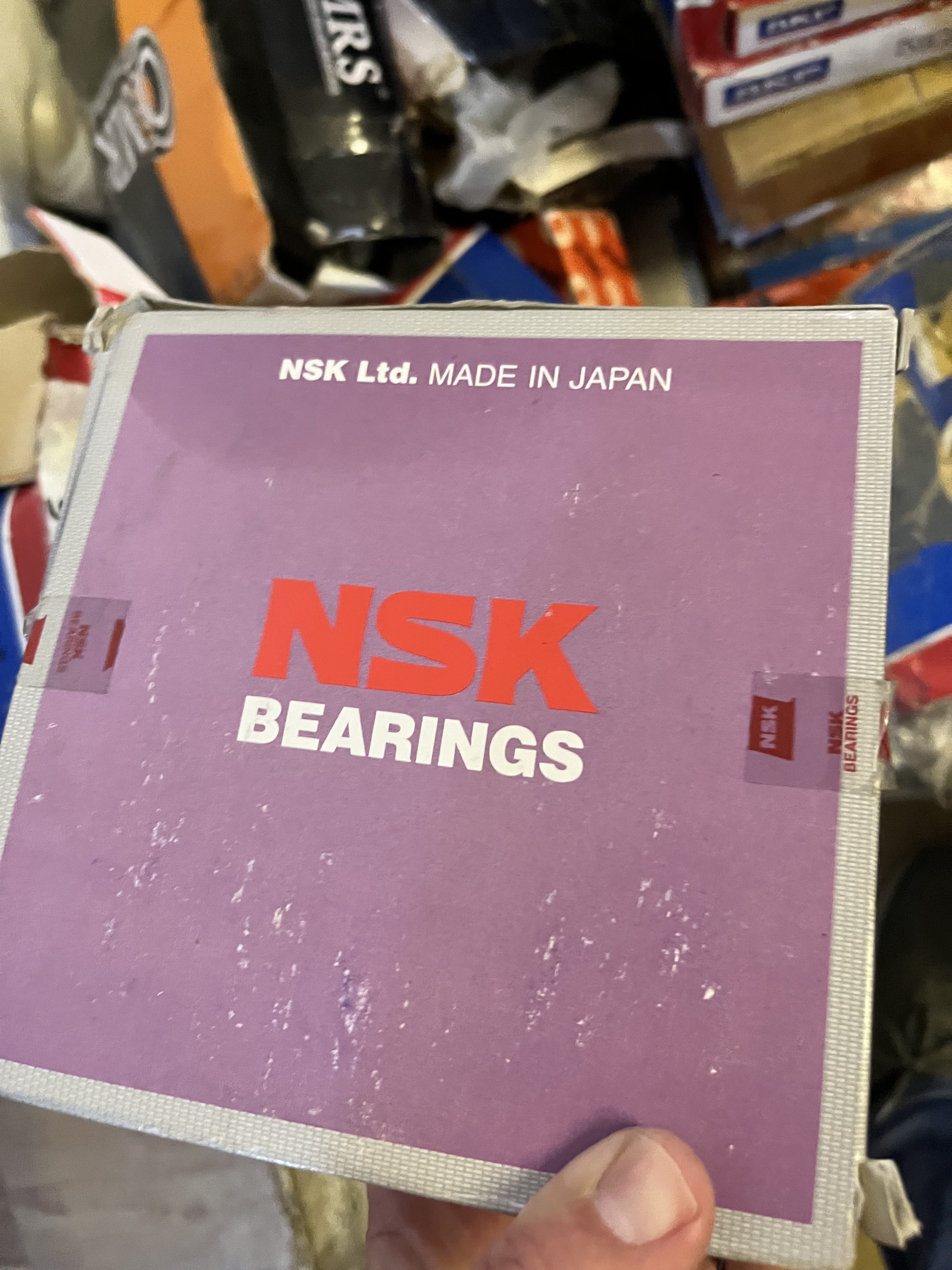 A person holds a used purple box labeled NSK BEARINGS with NSK Ltd. MADE IN JAPAN at the top. The worn box, likely containing LM6000 spare parts, is surrounded by other boxes in the background.