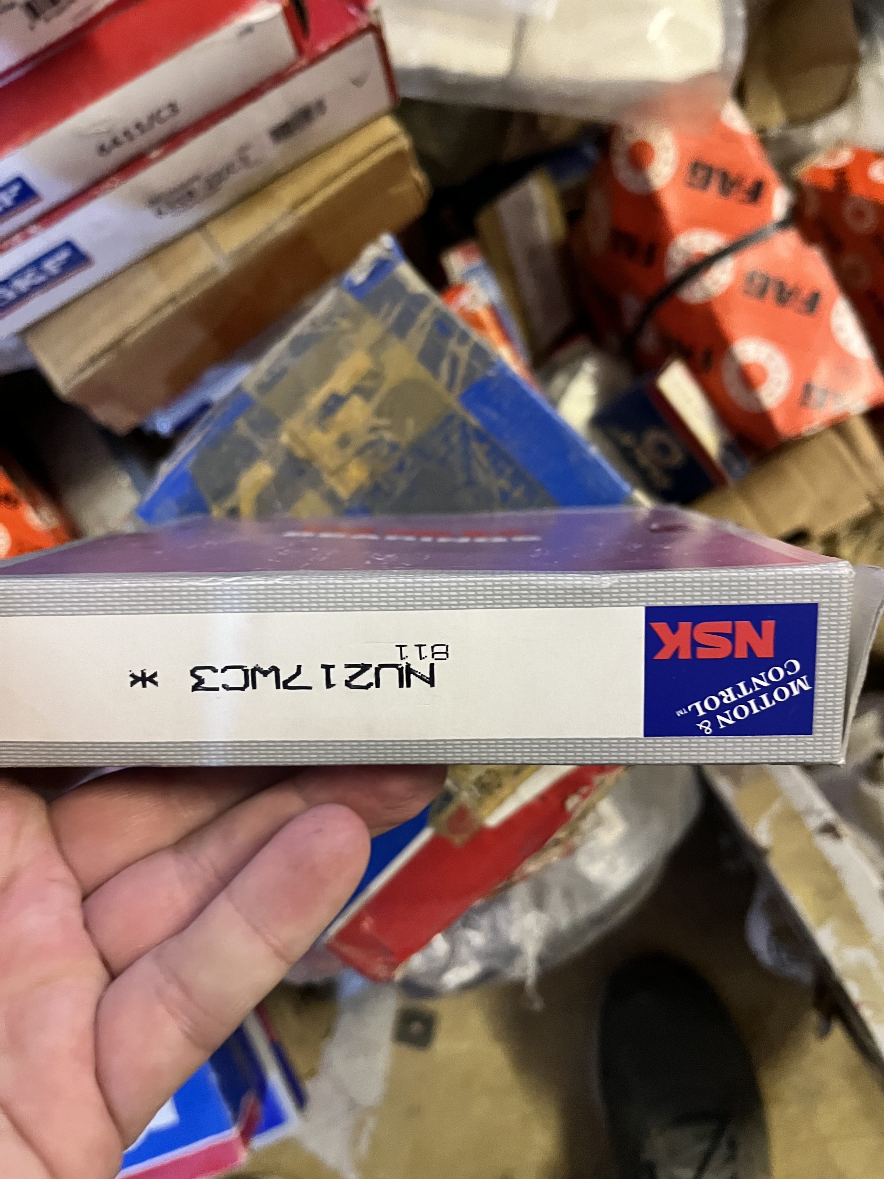 A hand holds an NSK bearing box upside down, displaying the label NU217MC3 8T1 Motion & Control with other stacked boxes in the background, often used in refurbished gas turbine applications like the LM6000.