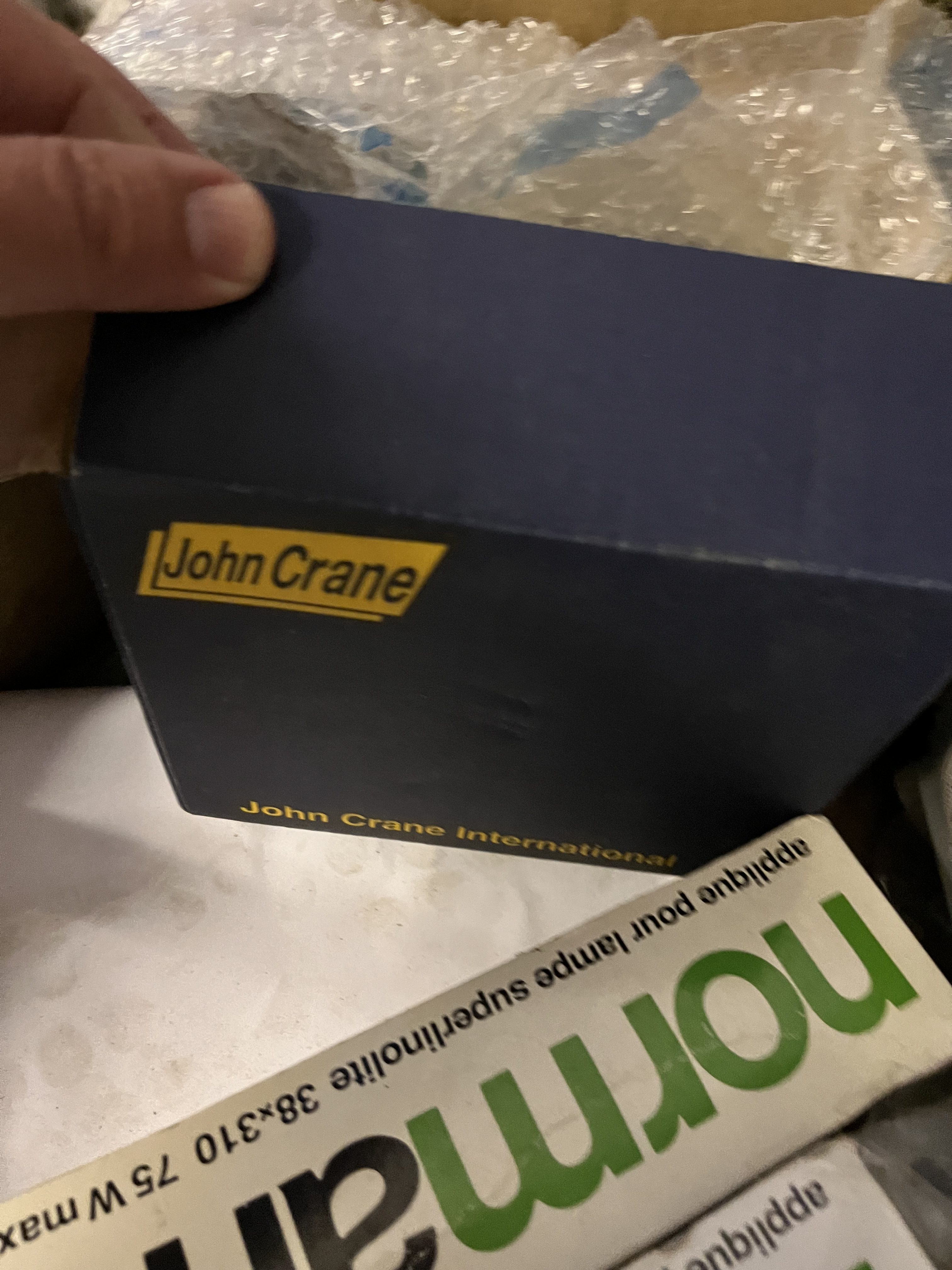 A hand holds a blue John Crane box with yellow text—possibly a refurbished part—partially out of a bubble wrap-filled container, next to another white and green NorMar package, used in gas turbine or obsolete equipment maintenance.