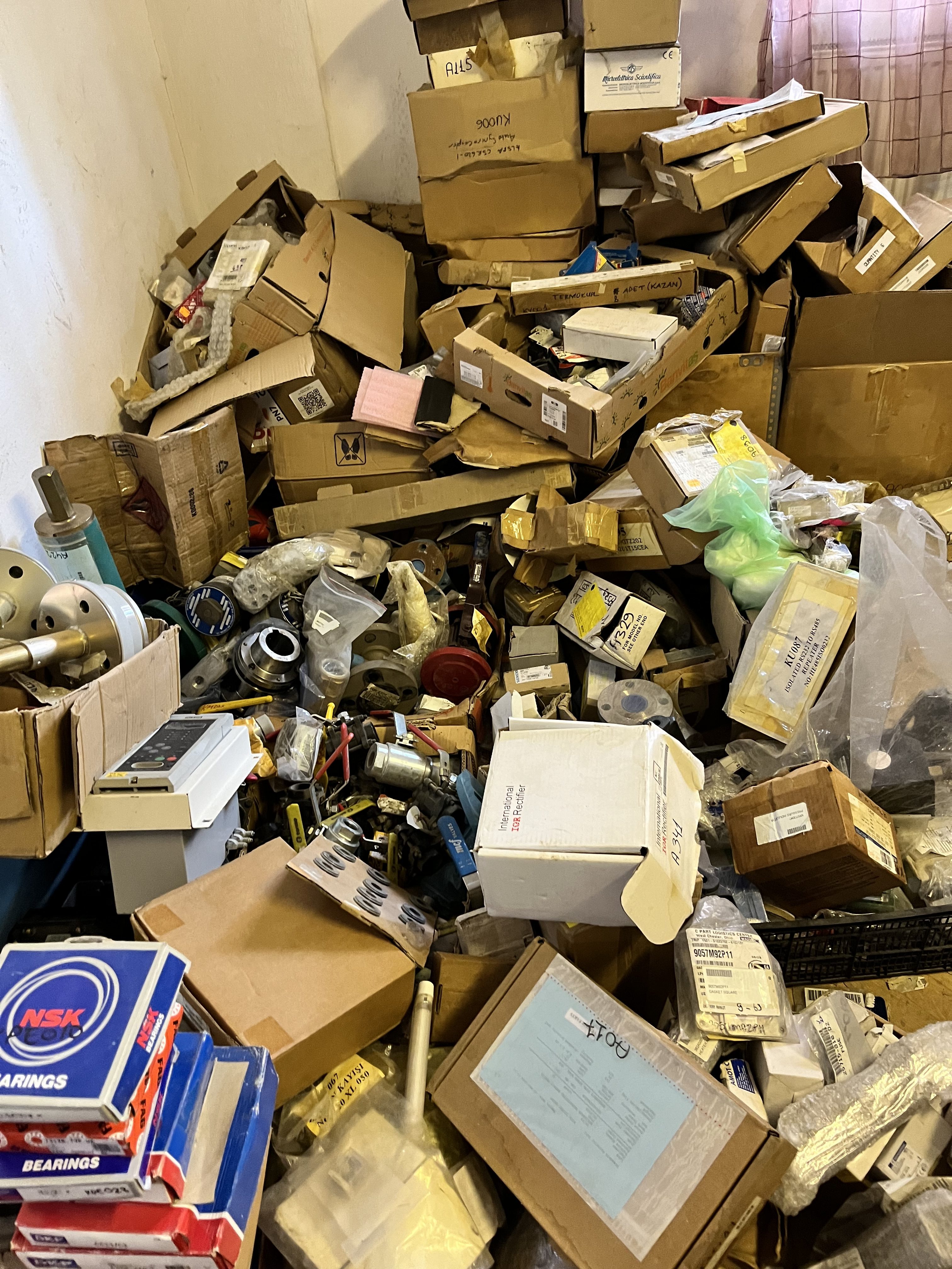 A cluttered pile of assorted boxes, used packages, and LM6000 gas turbine parts scattered on the floor and against a wall, with some labeled items and various tools visible among the disorganized heap.
