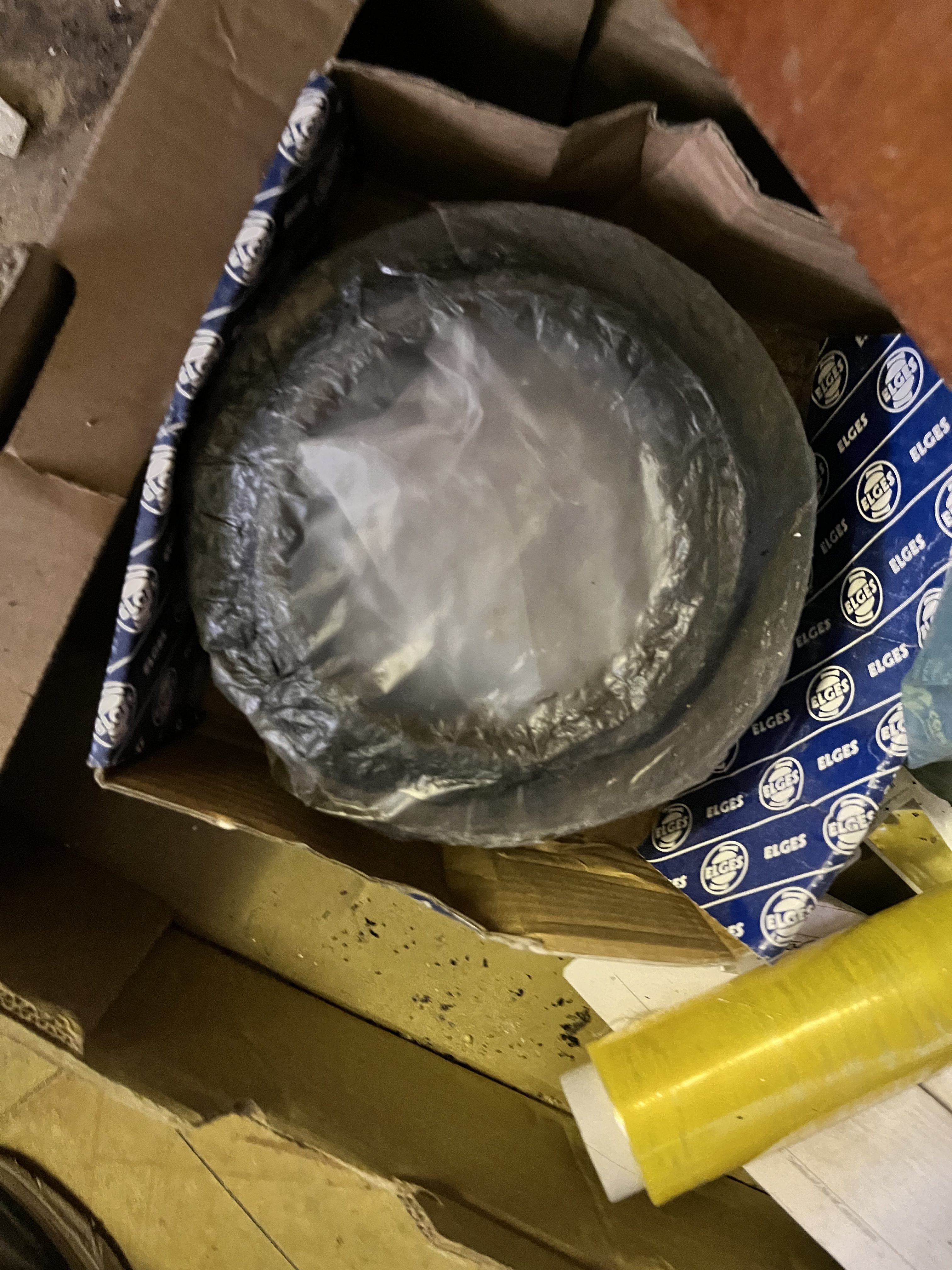 A large, round metal bearing—possibly refurbished—wrapped in plastic sits inside an open cardboard box lined with blue and white ELGES branded paper. The box, labeled LM6000, rests on a cluttered surface with yellow plastic wrap nearby.