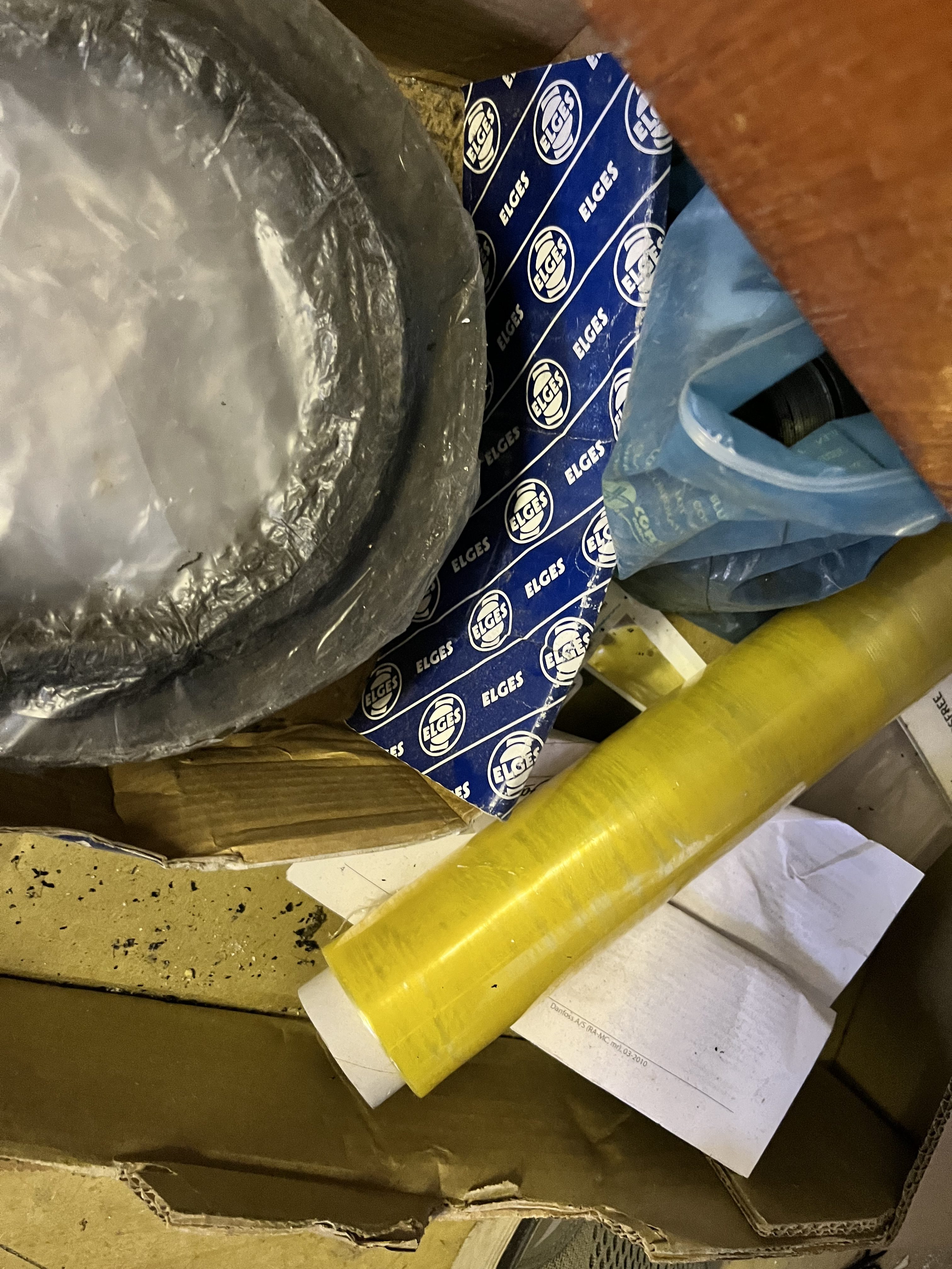 A close-up of a cluttered box containing obsolete spare parts: a plastic-wrapped black ring, a blue and white ELGES branded wrapper, a yellow plastic roll, papers, and other miscellaneous items.