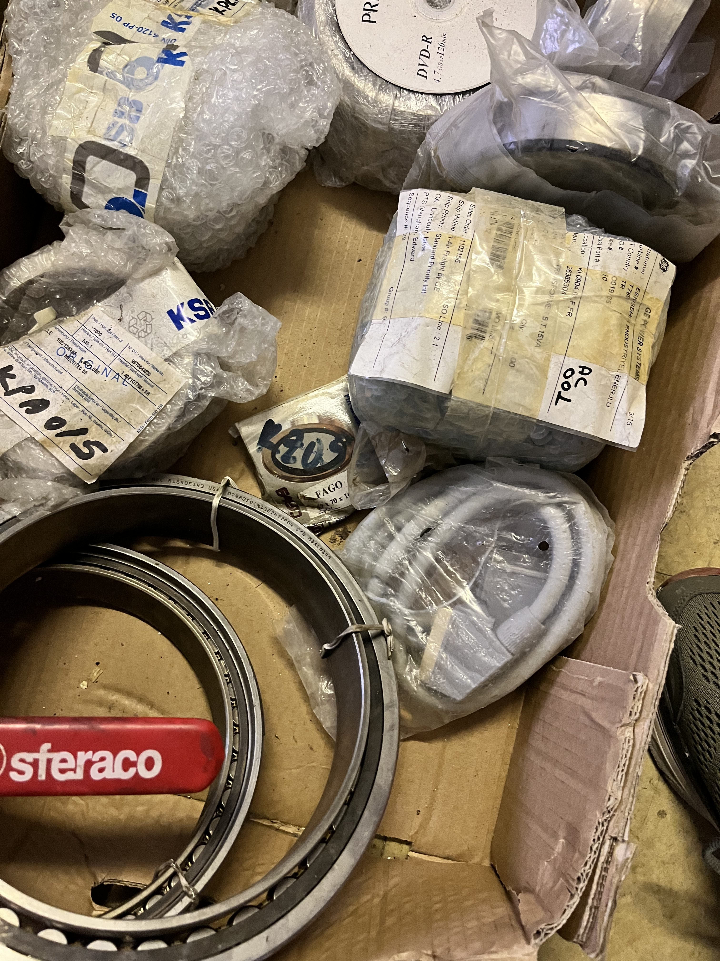 A cardboard box containing various industrial parts, including sealed bearings, refurbished components, coiled cables, and a red item labeled sferaco. Some potentially obsolete parts are wrapped in bubble wrap and plastic packaging.