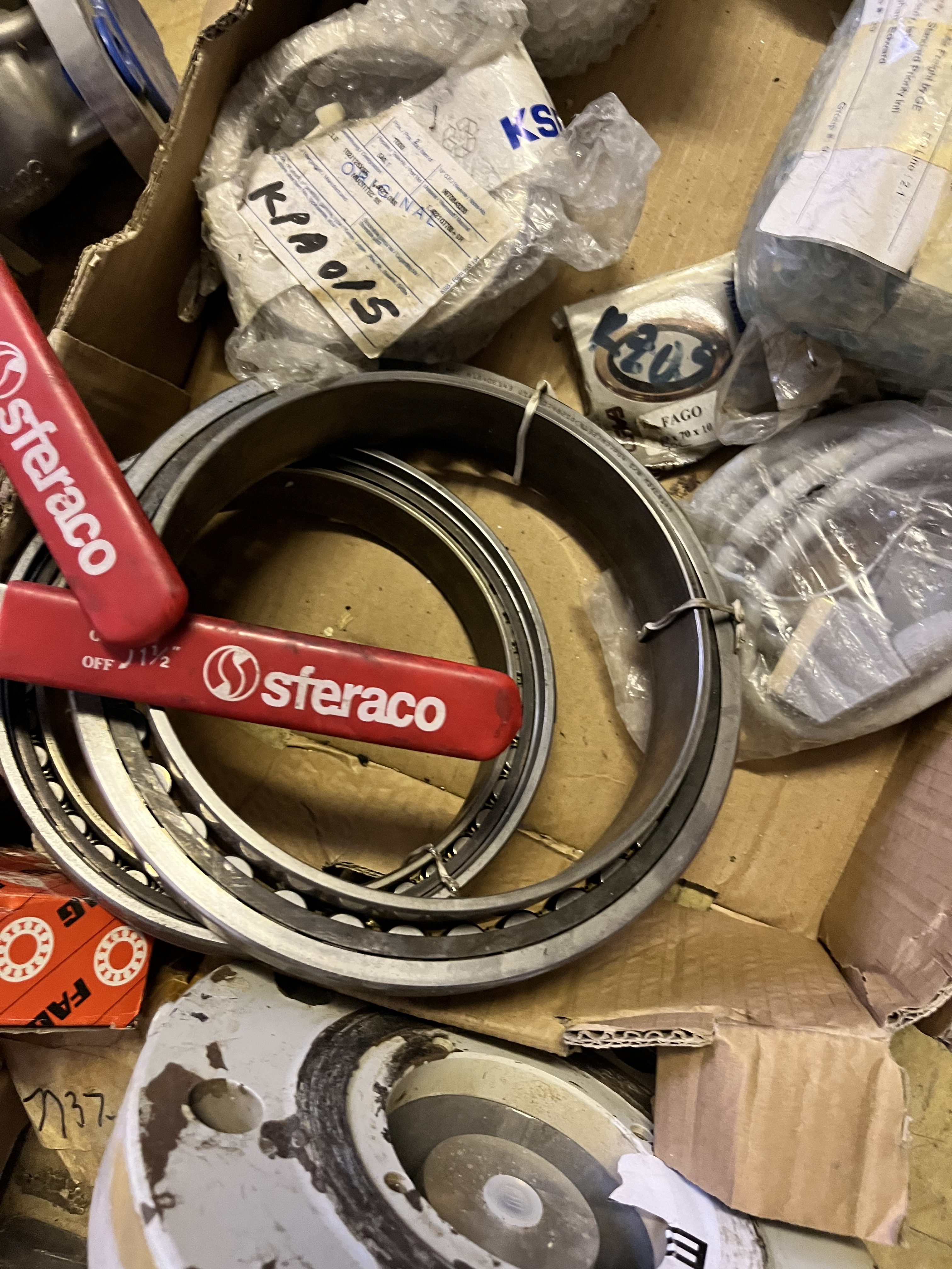 A close-up of assorted refurbished mechanical parts in a cardboard box, including metallic rings, a red lever labeled sferaco, wrapped components, and various obsolete bearings and industrial hardware.