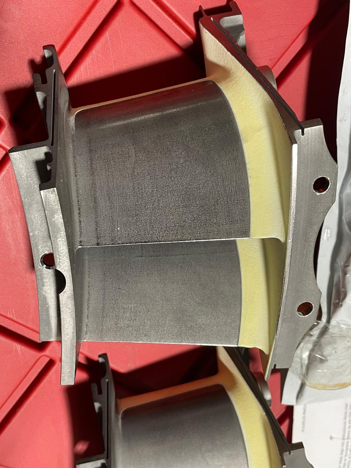 Three new surplus GE 2080M12G07 nozzles with metallic, curved blades and yellow edges rest on a red surface. Light highlights their smooth contours and precise construction as high-quality spare parts.