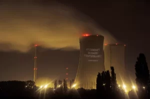 Night view of a nuclear power plant with cooling towers Steam rises as silhouettes of trees and the obsolete facility are visible in the foreground.