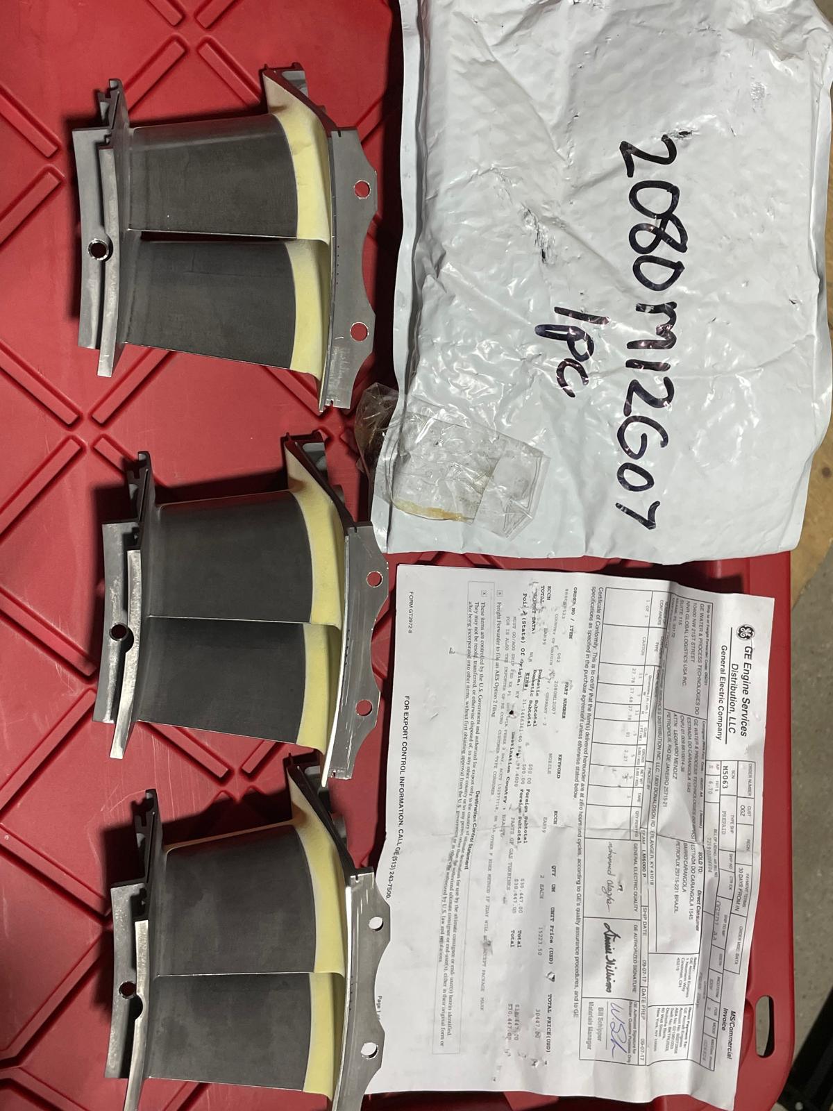 Three available nozzle GE 2080M12G07 new surplus pieces are laid out on a red surface next to a white shipping envelope labeled 208DM12607 1PC and a printed document with text and signatures.