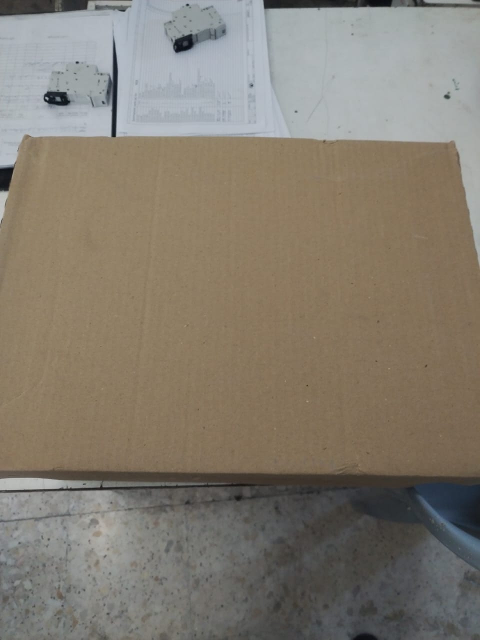 A large closed cardboard box, possibly containing Sold 3 pcs OBSOLETE GE FENRD/5 Door Mounted Extended Rotary Handle for GE Circuit Breaker, sits on a table with documents and two small electronic components in the background. The area shows signs of wear.