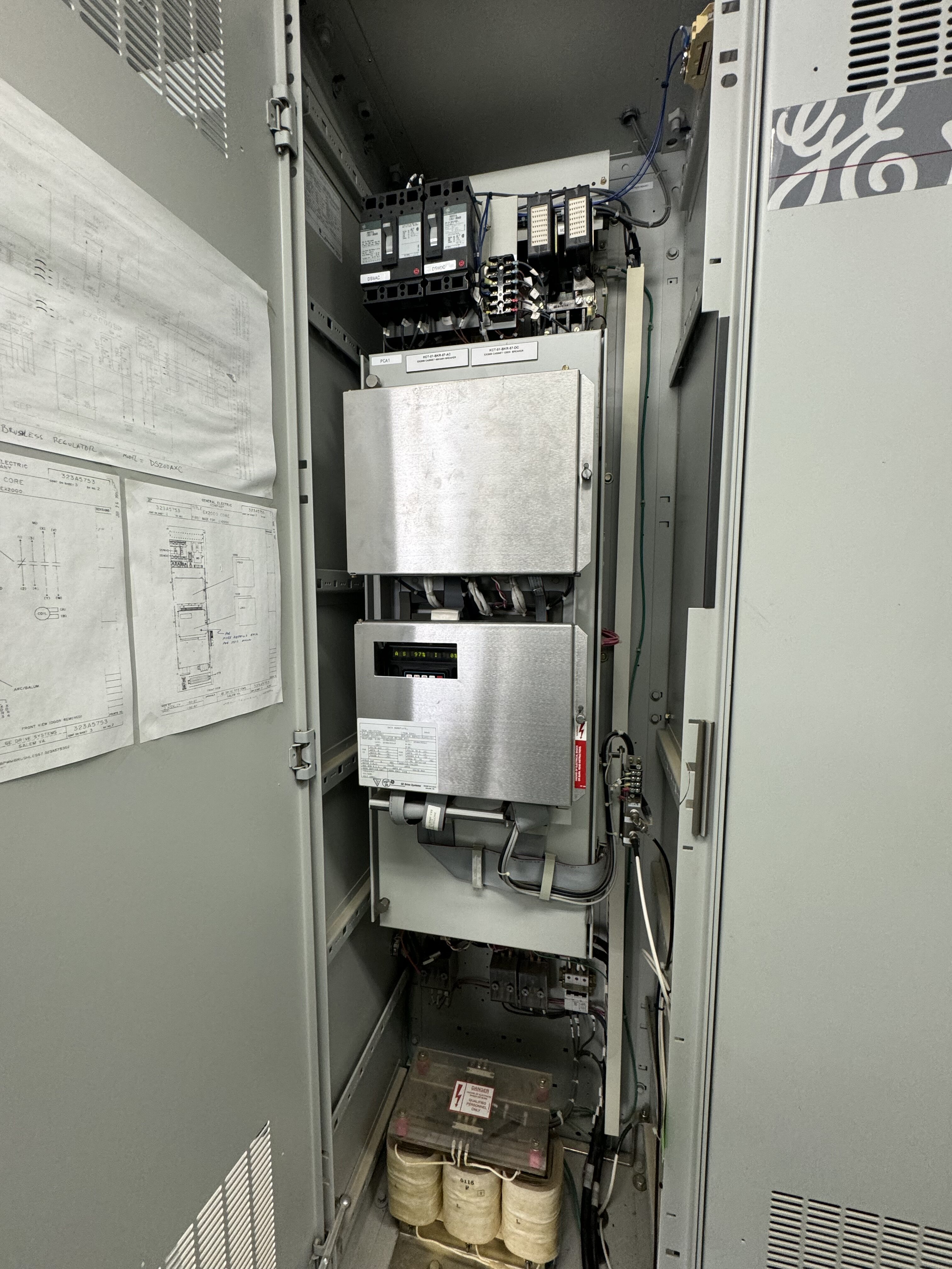 An open AVAILABLE MARK IV AND MARK V CONTROLS 01 industrial electrical cabinet shows circuit breakers, wiring, metal enclosures, technical diagrams on the door, and batteries at the bottom; the setup looks organized and well maintained.