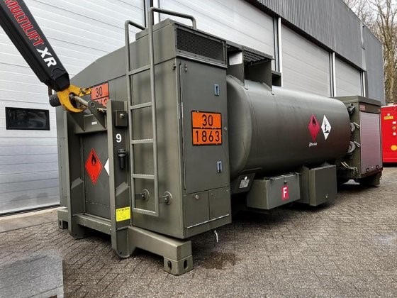 An olive-green Available Refueller Refueler Tank 10000 m3 with hazard signs is parked by gray garage doors, a crane arm on the left. This large tank is ready for heavy-duty fueling operations.