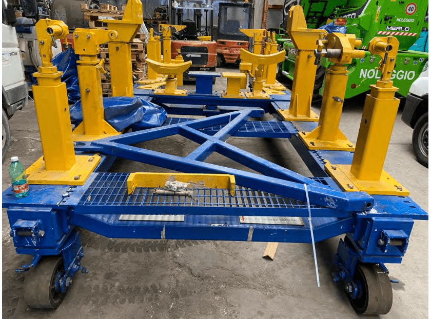 A large blue and yellow industrial metal frame with adjustable supports and wheels is stationed in a warehouse. Various machinery and equipment are visible in the background, along with a green vehicle marked NOLEGGIO.