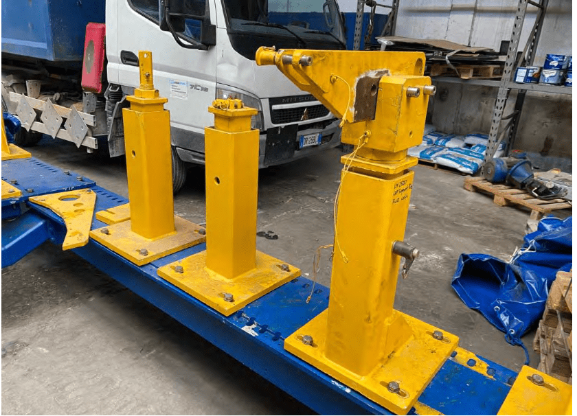 Three yellow metal support columns, one with an angled arm, mounted on a blue steel base in an industrial warehouse with a white truck and various materials in the background.