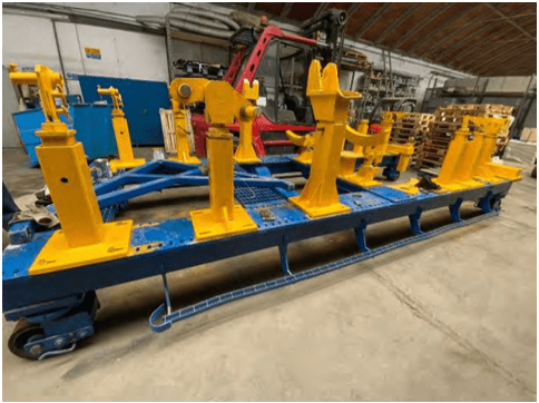 A large blue and yellow metal industrial fixture is positioned inside a spacious warehouse with a concrete floor; shelves and stacked pallets are visible in the background. Stand for gas turbine LM6000 and LM2500