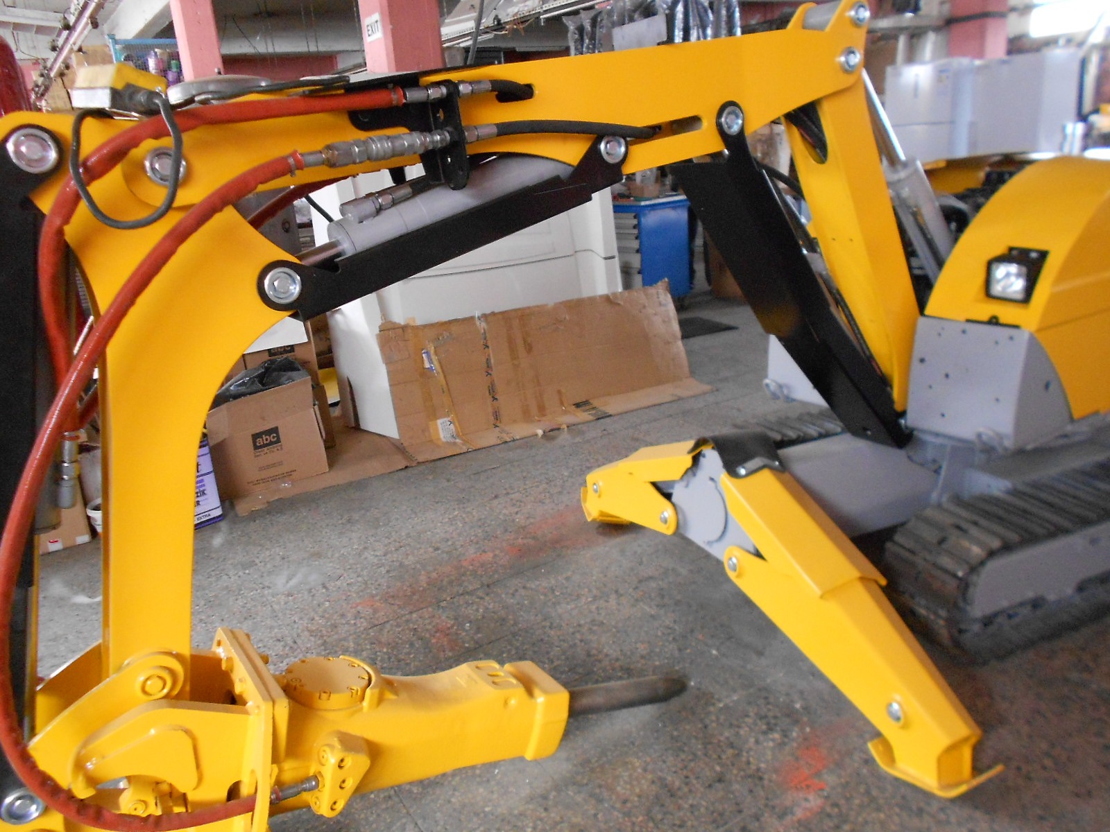 Close-up of the DEMOLISHING UNIT BROKK 180 REV-F1’s yellow arm inside a warehouse, highlighting hydraulic hoses, bolts, and its gray body, with cardboard boxes and other industrial items seen in the background.