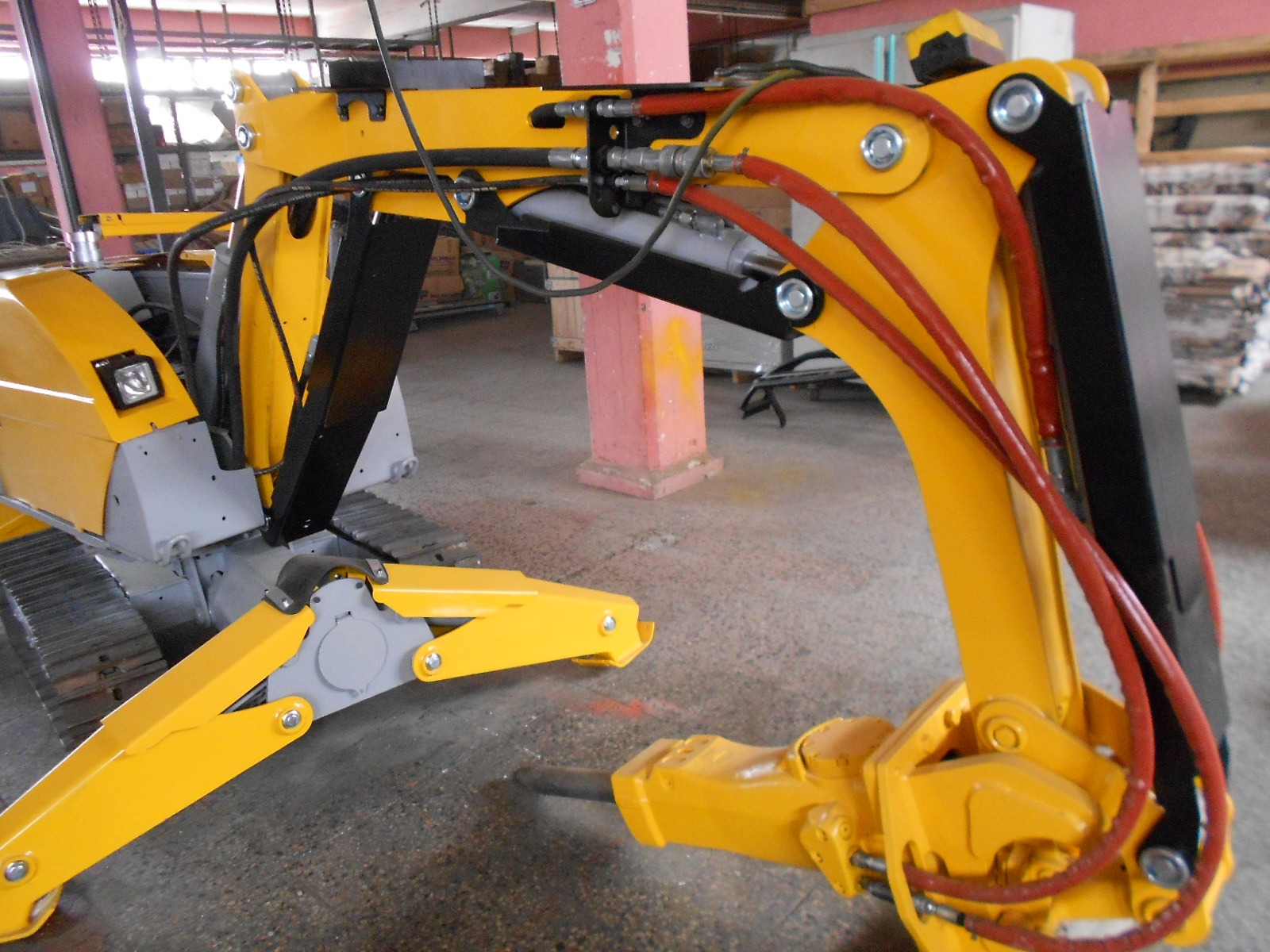 A close-up of the DEMOLISHING UNIT BROKK 180 REV-F1’s yellow hydraulic excavator arm indoors displays hoses, joints, and mechanical details, with part of its tracks and a pink support column visible in the background.