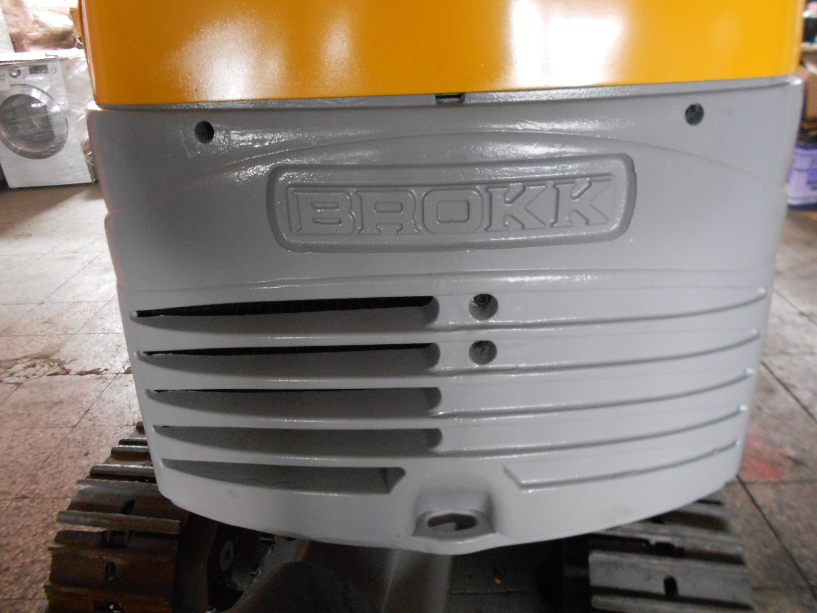 Close-up of the gray and yellow exterior of a DEMOLISHING UNIT BROKK 180 REV-F1, featuring the BROKK brand name embossed on the front panel above vented slats, with part of its rubber track visible below.