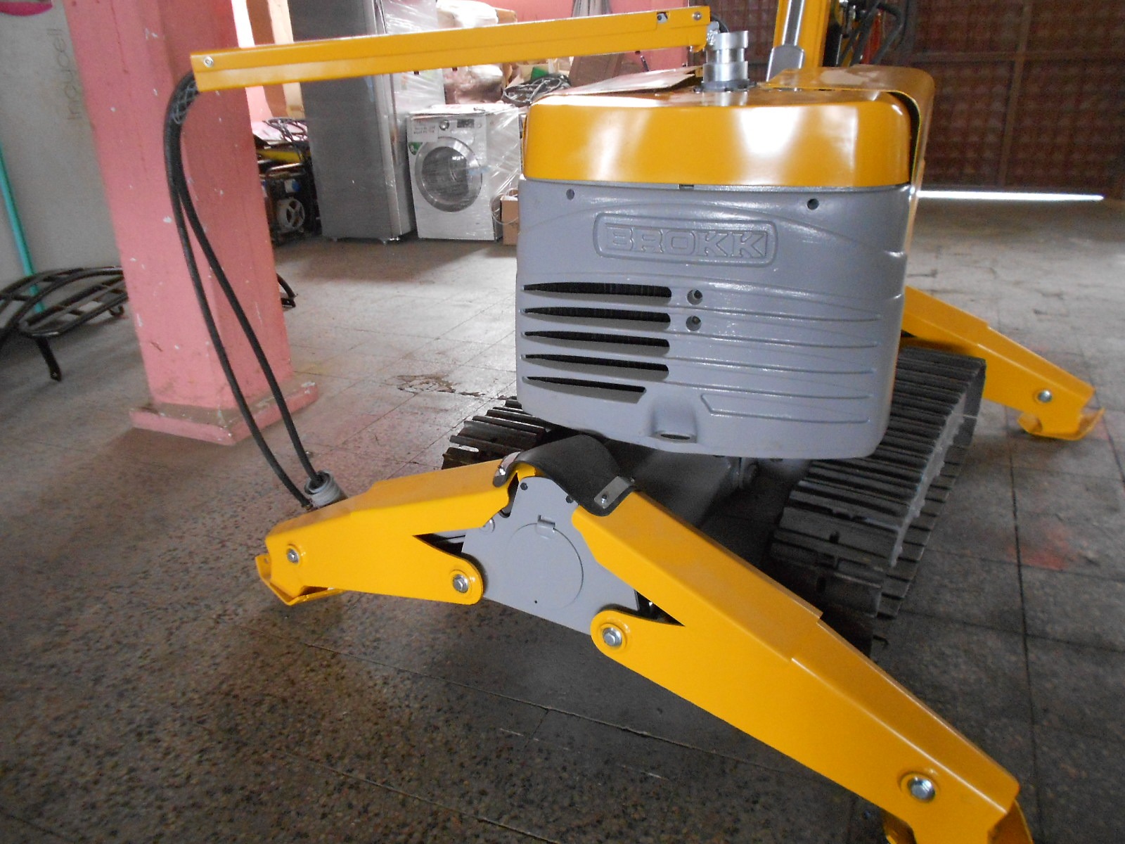A close-up of the DEMOLISHING UNIT BROKK 180 REV-F1, a yellow and gray machine with tank-like tracks, is seen positioned indoors on a tiled floor, likely used for construction or demolition tasks.