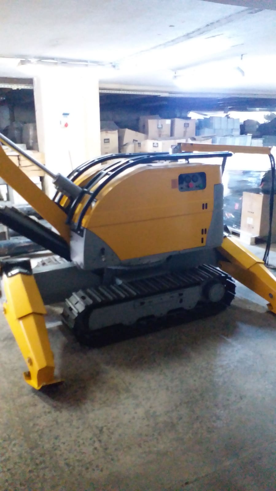 A yellow and gray DEMOLISHING UNIT BROKK 180 REV-F1 compact tracked demolition robot with hydraulic arms is parked indoors on a concrete floor, surrounded by cardboard boxes and shelves in a storage area.