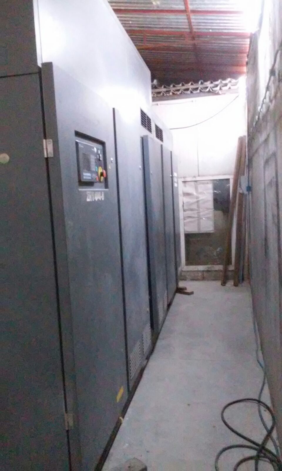 A narrow industrial room features large gray ATLAS COPCO ZH 10000-6-7PL AIR COMPRESSOR cabinets on the left, a concrete floor, a white window at the far end, and cables plus wooden planks leaning against the right wall.
