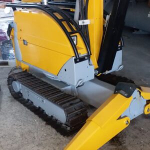 A yellow and gray DEMOLISHING UNIT BROKK 180 REV-F1 tracked excavator is parked indoors on a concrete floor, its arm partially extended with a cardboard box resting on top.
