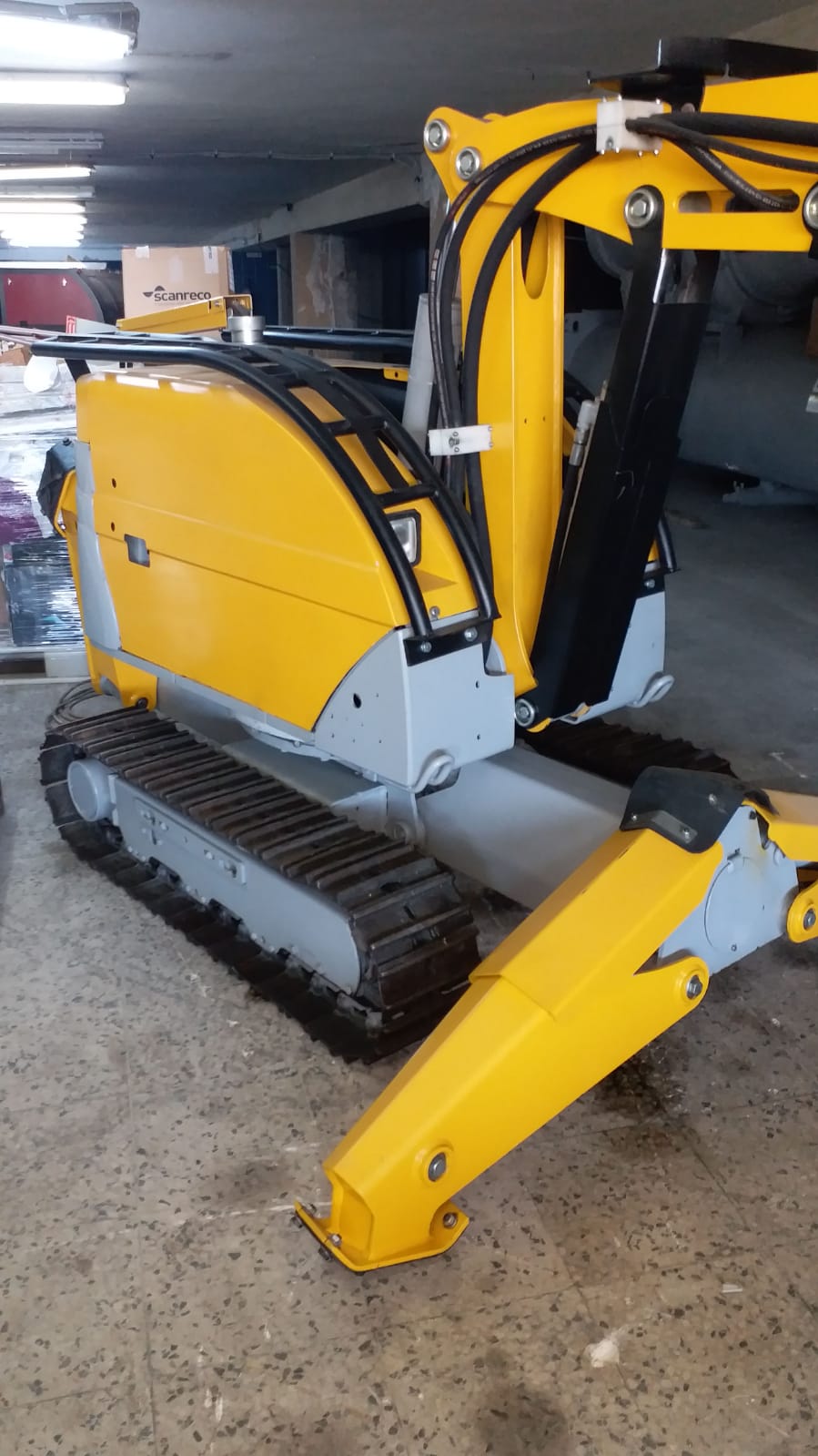 A yellow and gray DEMOLISHING UNIT BROKK 180 REV-F1 tracked excavator is parked indoors on a concrete floor, its arm partially extended with a cardboard box resting on top.